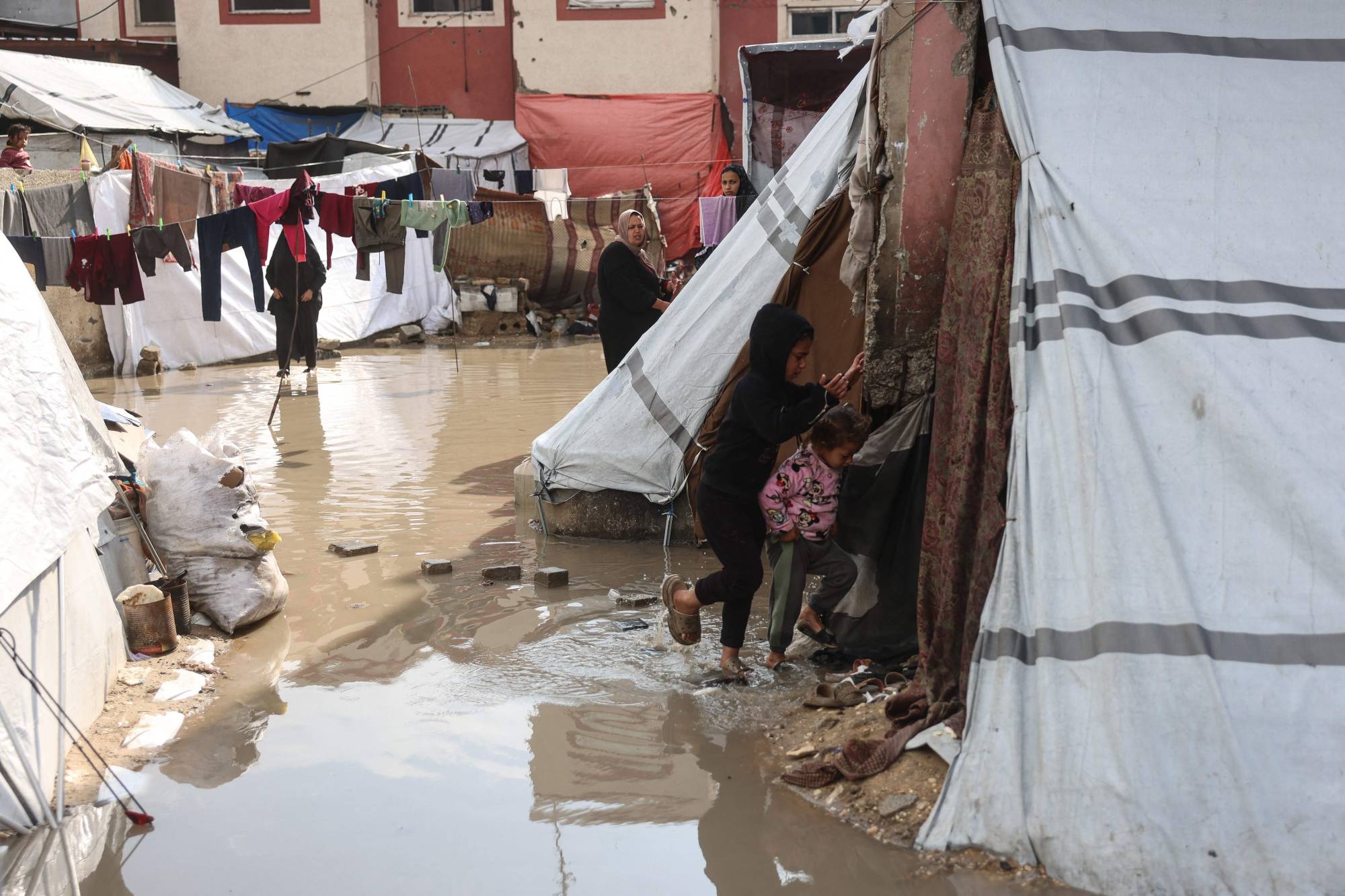 Güney Gazze Şeridi'ndeki Han Yunus'ta, yerinden edilmiş Filistinlilerin barındığı derme çatma bir kampta, çocuklar sular altında kalmış bir sokaktan geçiyor (AFP)