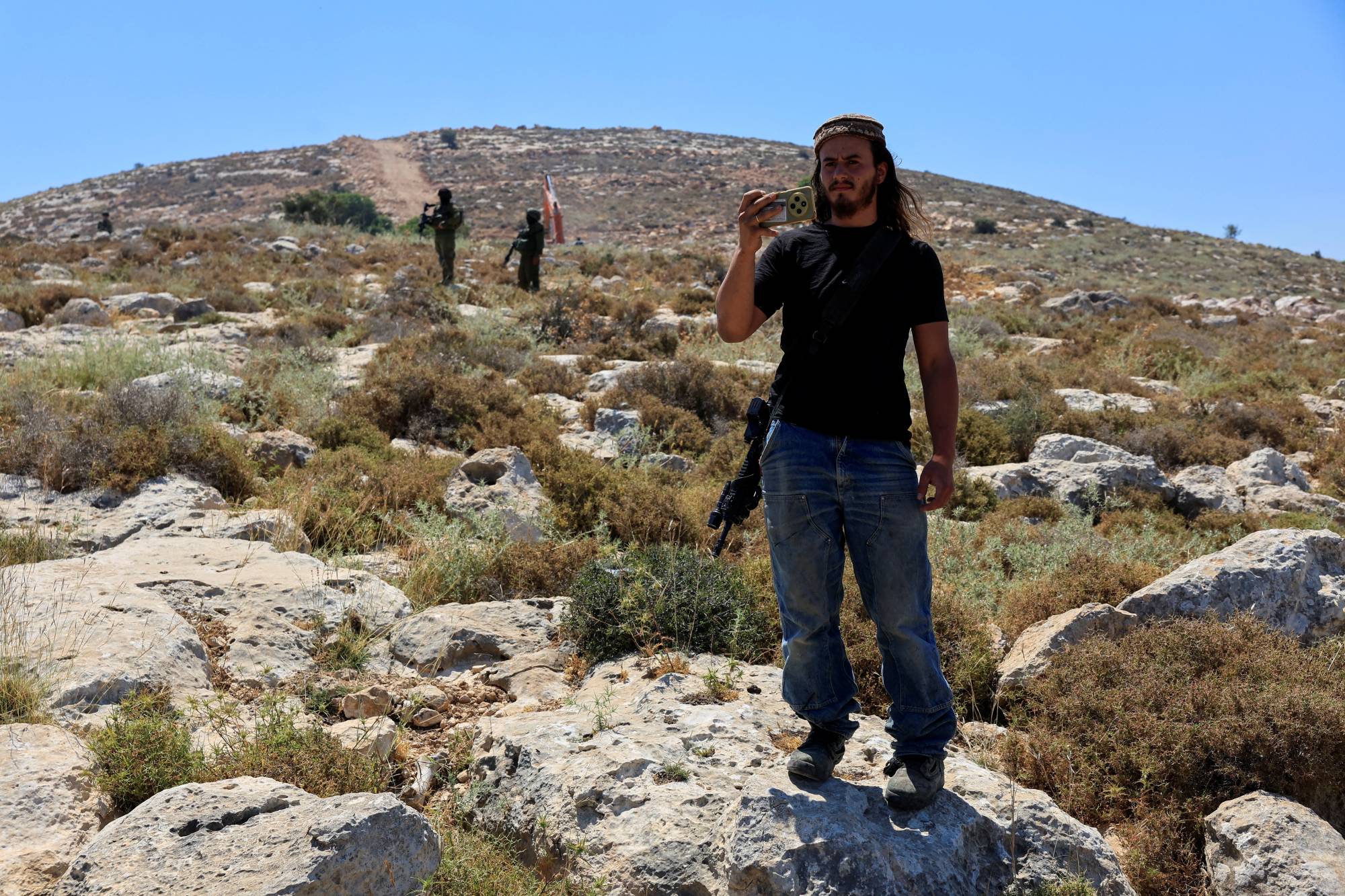 Batı Şeria’nın El-Halil kenti yakınlarındaki bir yerleşim birimine karşı düzenlenen protesto esnasında, İsrailli askerler nöbet tutarken bir İsrailli yerleşimci cep telefonuyla fotoğraf çekiyor, 9 Haziran 2026. (Reuters)