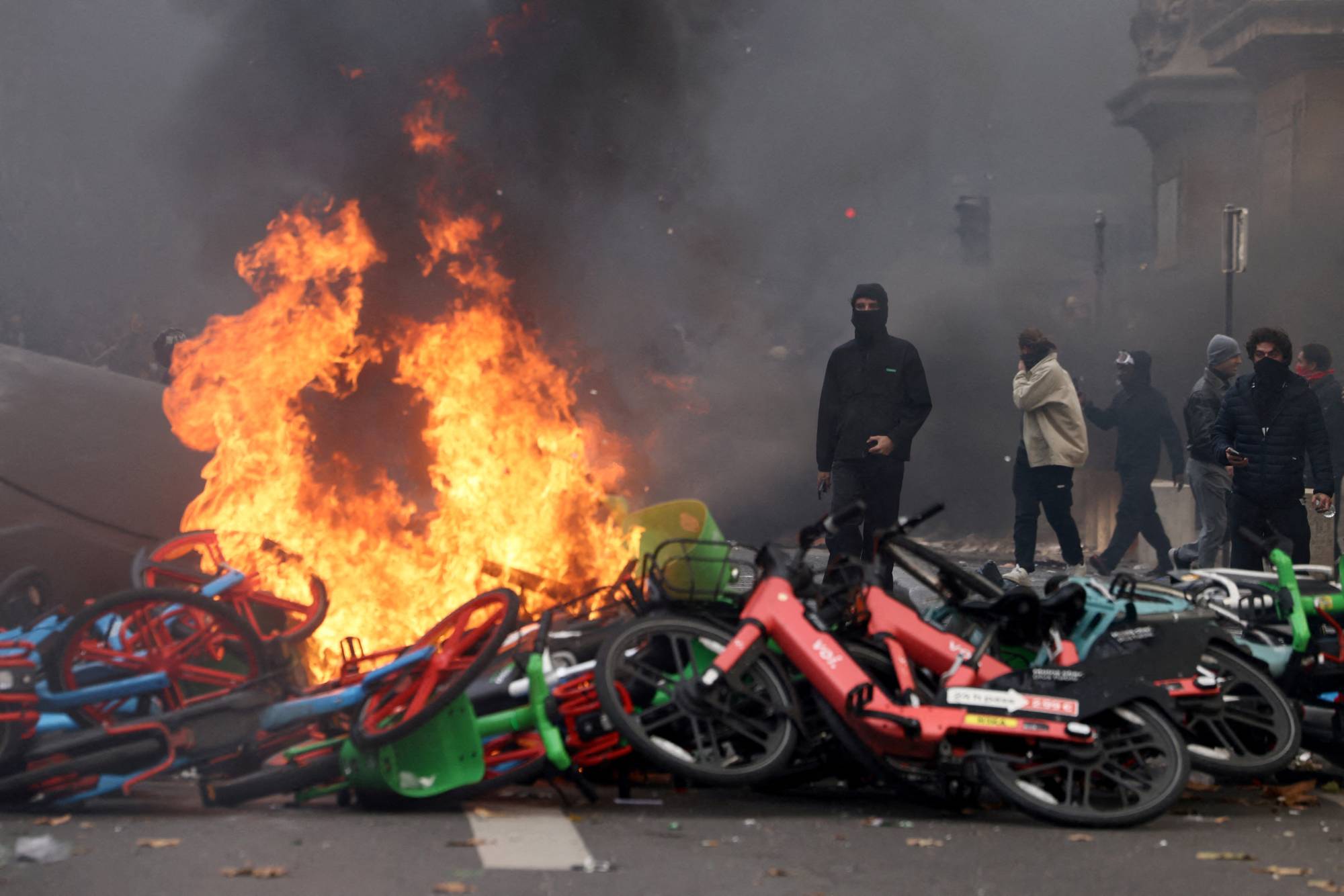 Paris’teki Cumhuriyet Meydanı’nda (Place de la République) yanan bisikletlerin yanında duran maskeli protestocular (Reuters)