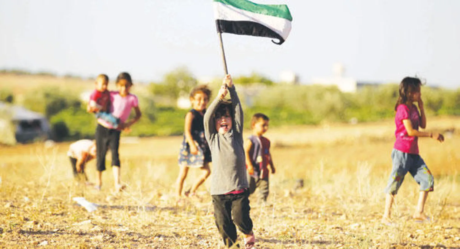أطفال سوريون يلهون ويرفع أحدهم علم المعارضة السورية في جنوب اسم (رويترز)
