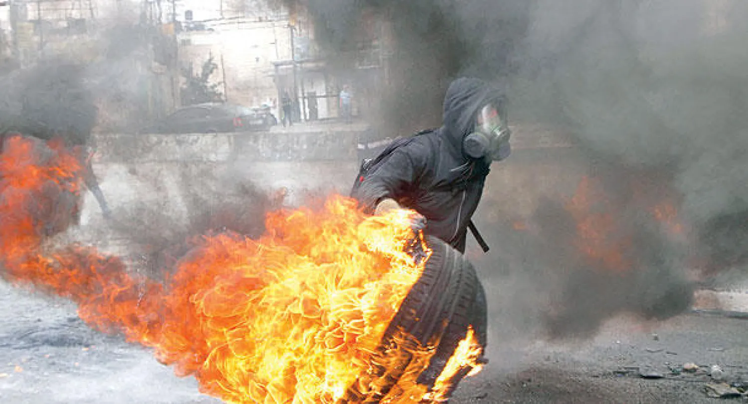 متظاهر فلسطيني مقنع يحمل إطار سيارة خلال تصادم مع الجيش الإسرائيلي في مخيم قلنديا القريب من مدينة رام الله أمس (رويترز)