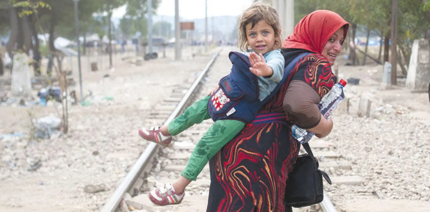 على الحدود بين اليونان ومقدونيا أمس لاجئة تحمل ابنتها على ظهرها تعبر سكة حديدية في بلدة جيفجيليجا (أ.ف.ب)