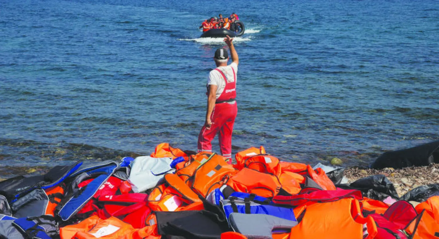 قارب مكتظ بالمهاجرين يصل إلى جزيرة ليزبوس اليونانية، بينما يلوح لهم أحد المواطنين اليونانيين وهو يقف إلى جانب مجموعة من سترات النجاة أمس (أ.ب)