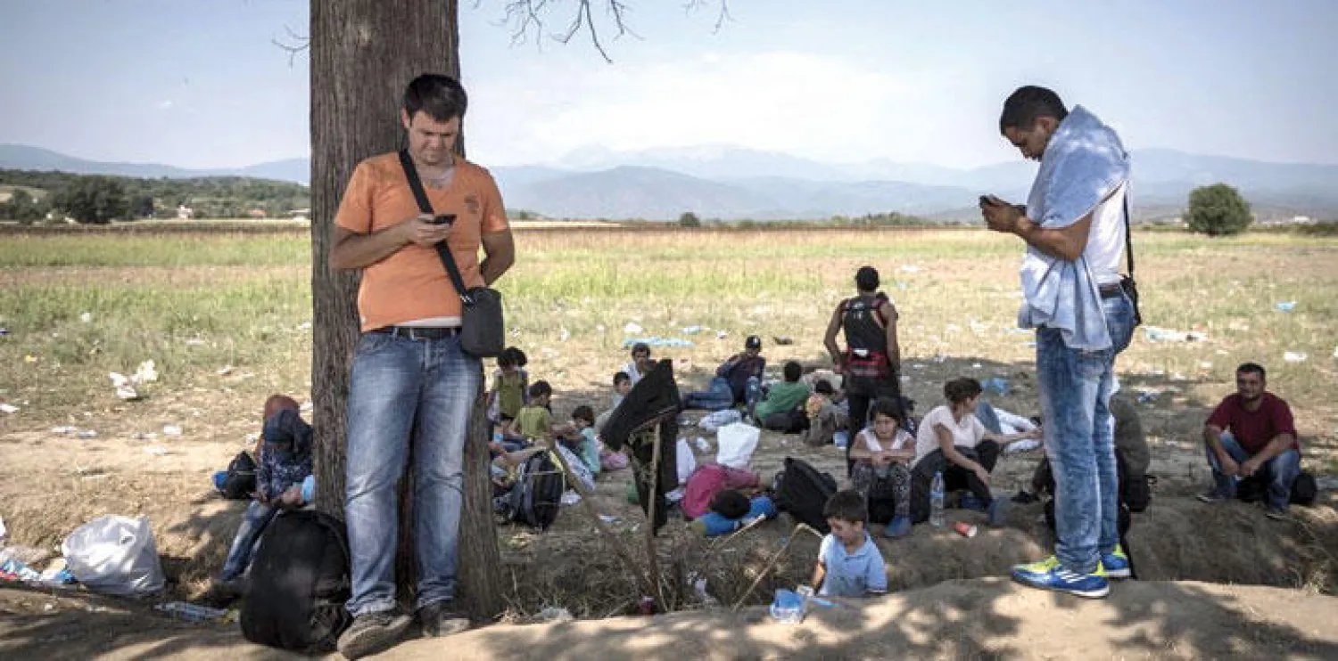 أحمد ماجد يتفقد هاتفه الجوال فيما يجلس أفراد عائلته ولاجئون آخرون أرضًا عند الحدود المقدونية - اليونانية، في 26 أغسطس (آب) الماضي («نيويورك تايمز»)