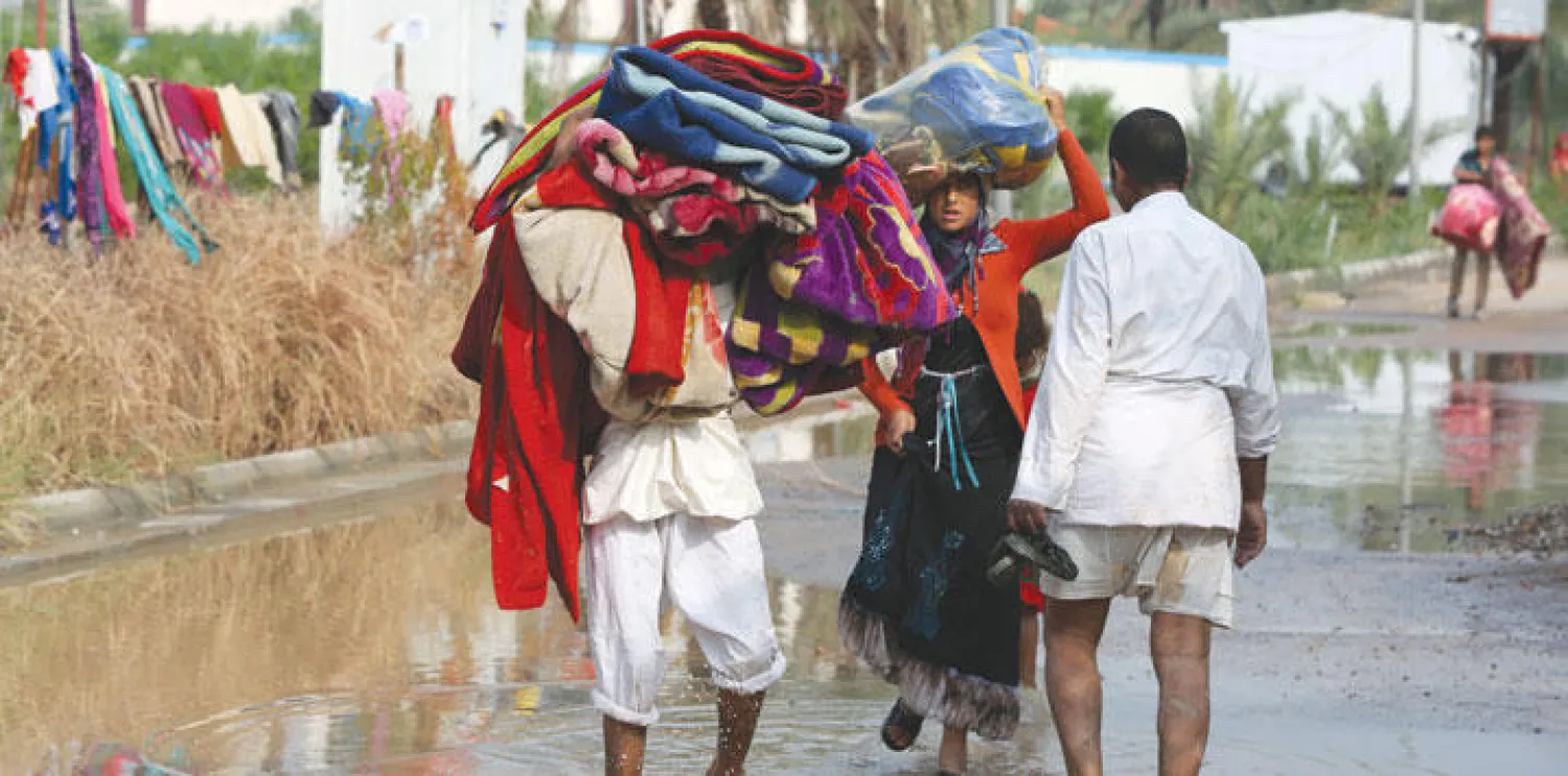نازحون يحملون فرشًا وبطانيات وسط مياه الأمطار في مخيم للنازحين قرب بغداد أمس (أ.ف.ب)