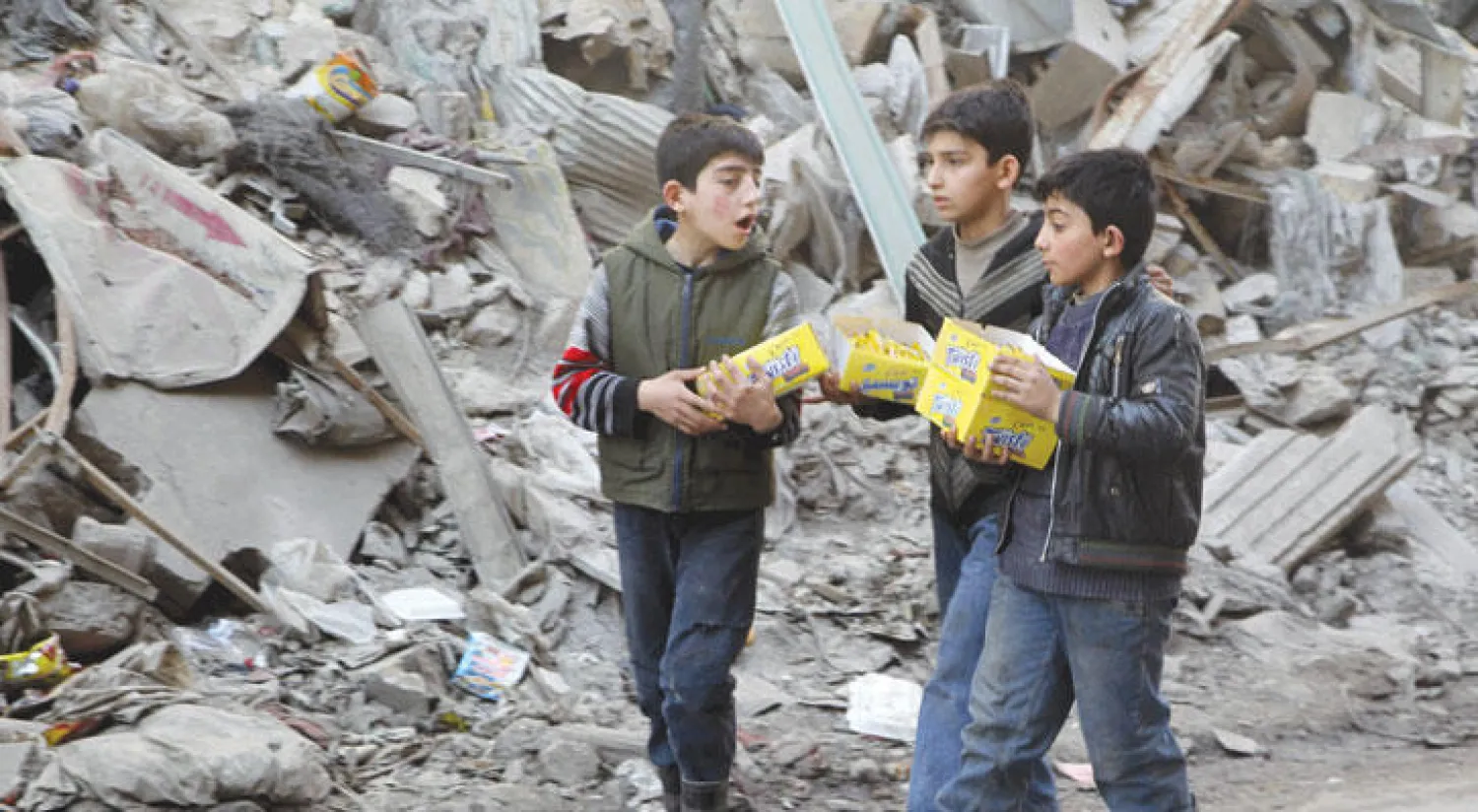 أطفال بريف حلب الشمالي يحملون علب بسكويت قرب ركام الأبنية المدمرة بسبب غارات الطيران ليبيعوها للمارة (رويترز)