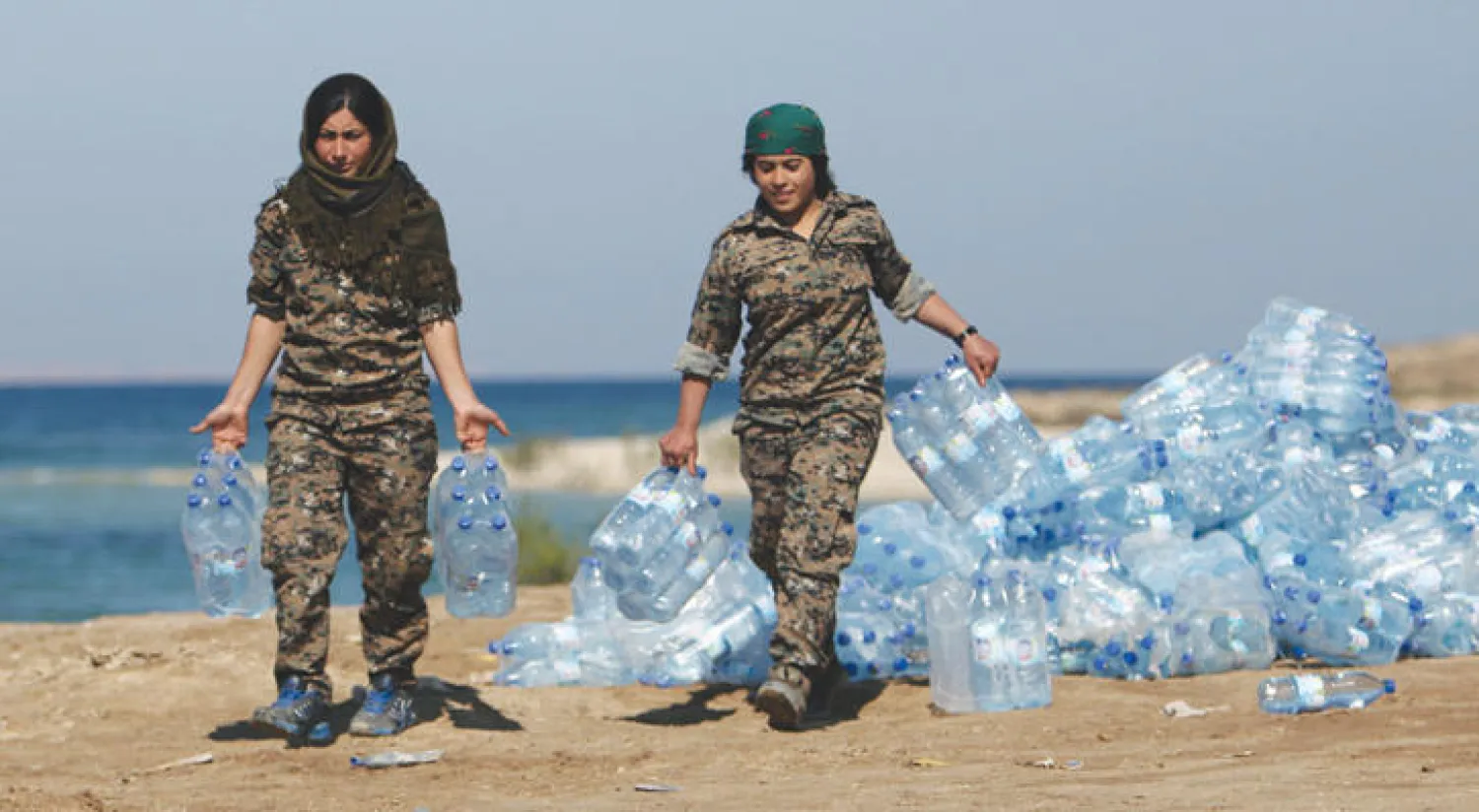 مقاتلتان من «قوات سوريا الديمقراطية» تحملان عبوات ماء بالقرب من نهر الفرات الذي تجري قرب ضفافه المعارك مع «داعش» (رويترز)