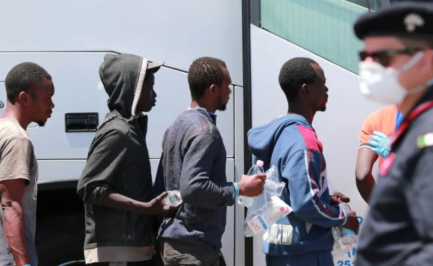 ضابط شرطة إيطالي يقف أمام مهاجرين لدى نزولهم من سفينة الحراسة المدنية الإسبانية سيغورا الإسبانية تم إنقاذهم في البحر الأبيض المتوسط (أ.ف.ب)