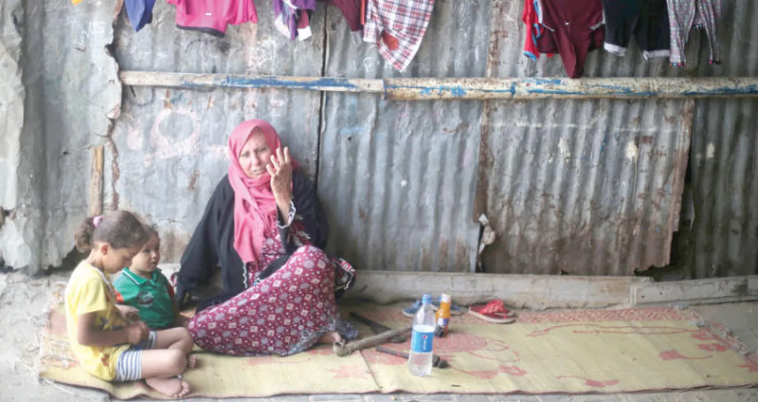 فلسطينية تجلس مع حفيديها قرب جدار بيتها الصفيحي بمخيم خان يونس جنوب قطاع غزة (رويترز)