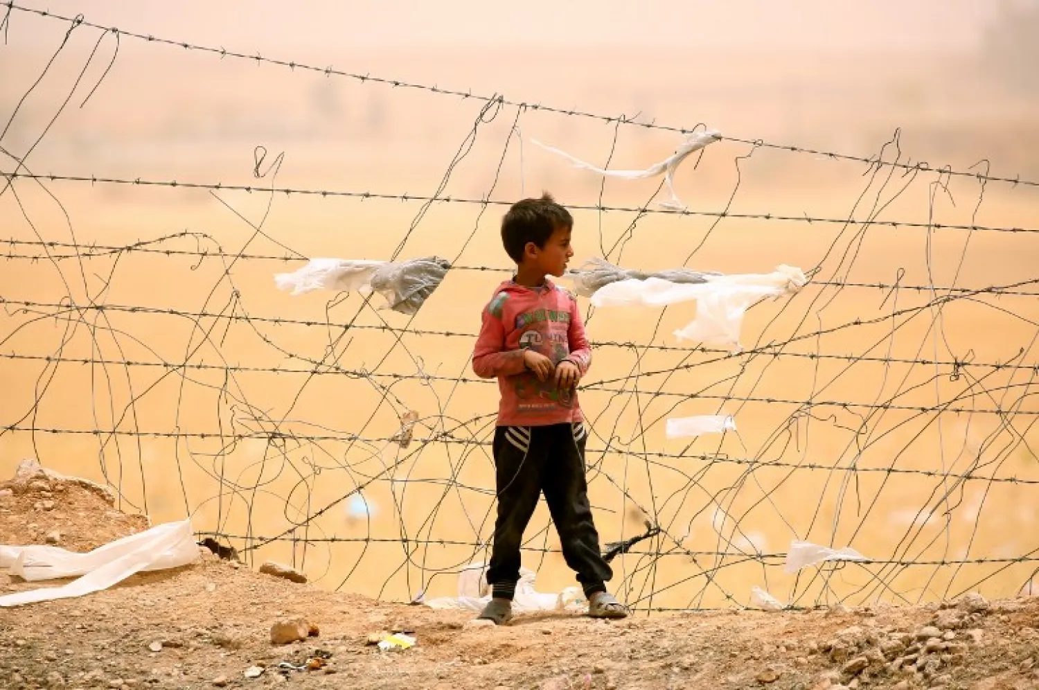 A displaced Syrian boy who fled the ISIS stronghold of Raqqa stands near a fence during a sandstorm at a temporary camp in the northern Syrian village of Ain Issa on May 19, 2017. / AFP PHOTO / DELIL SOULEIMAN