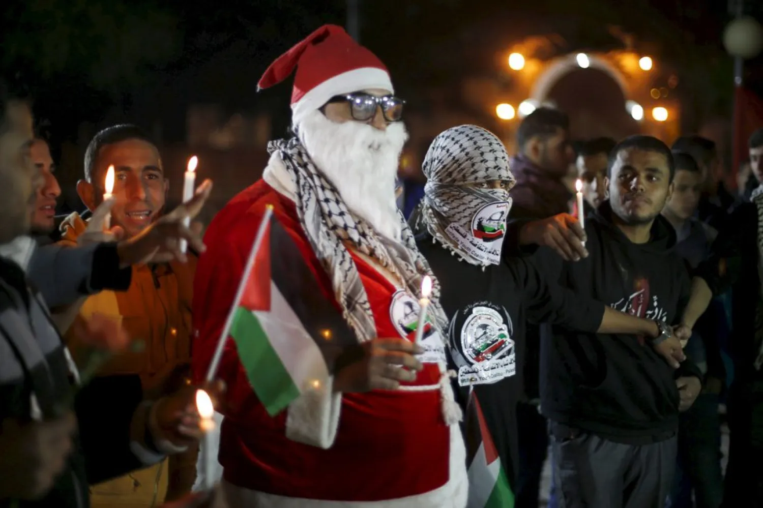 A masked Palestinian stands next to a man wearing a Santa Claus costume as they hold candles during a rally on Christmas day. (File Photo: Suhaib Salem, Reuters,12/26/2015)
