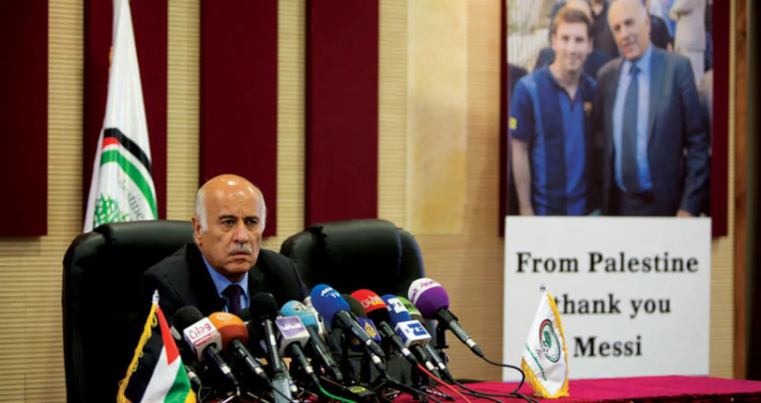 جبريل الرجوب يتحدث إلى الصحافيين ويظهر على جدار مكتبه إلى يساره صورة تجمعه بميسي (أ.ب)