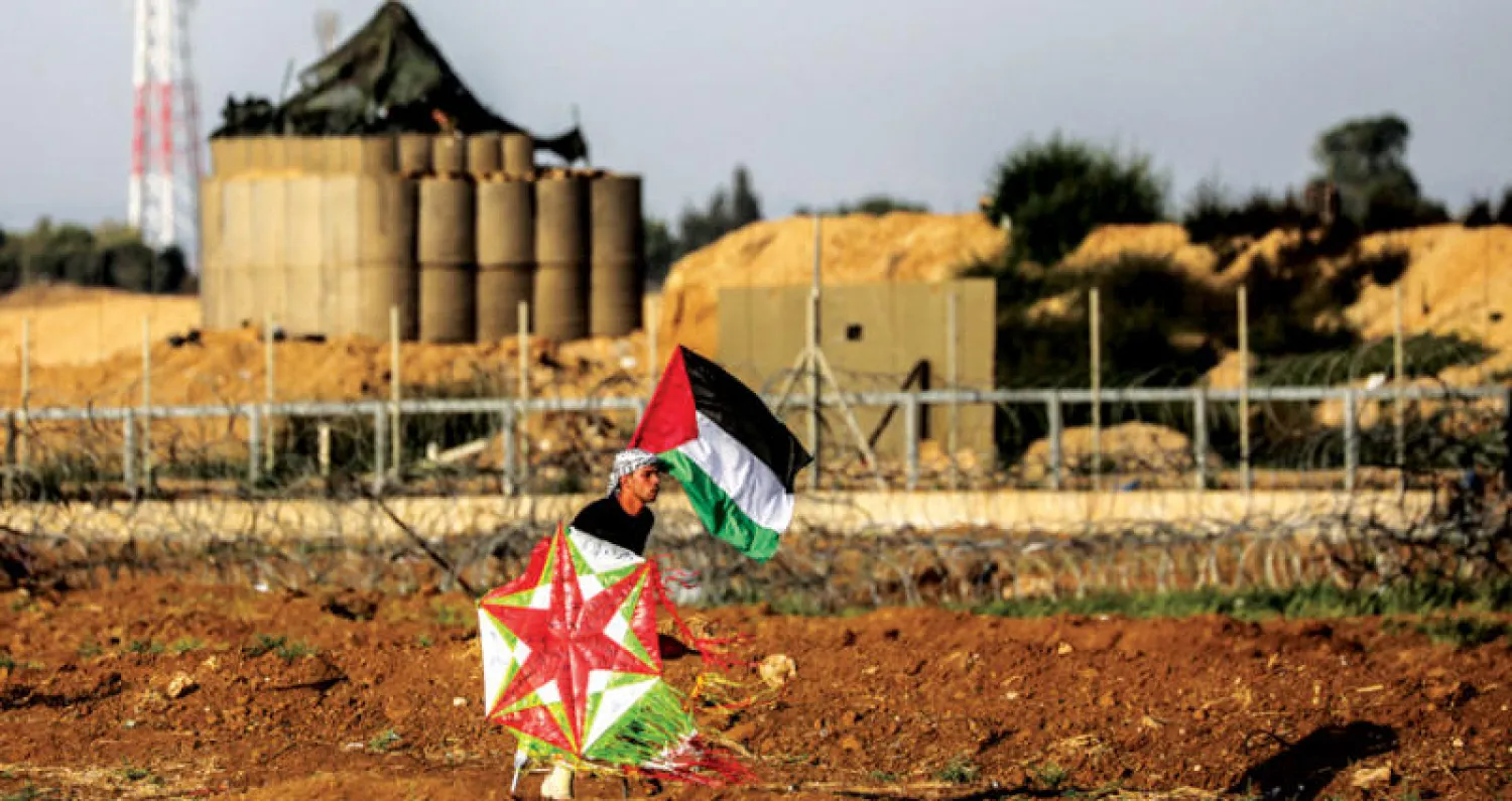 شاب يرفع العلم الفلسطيني بيد ويحمل طائرة ورقية بالأخرى خلال احتجاجات على حدود غزة (إ.ب.أ)