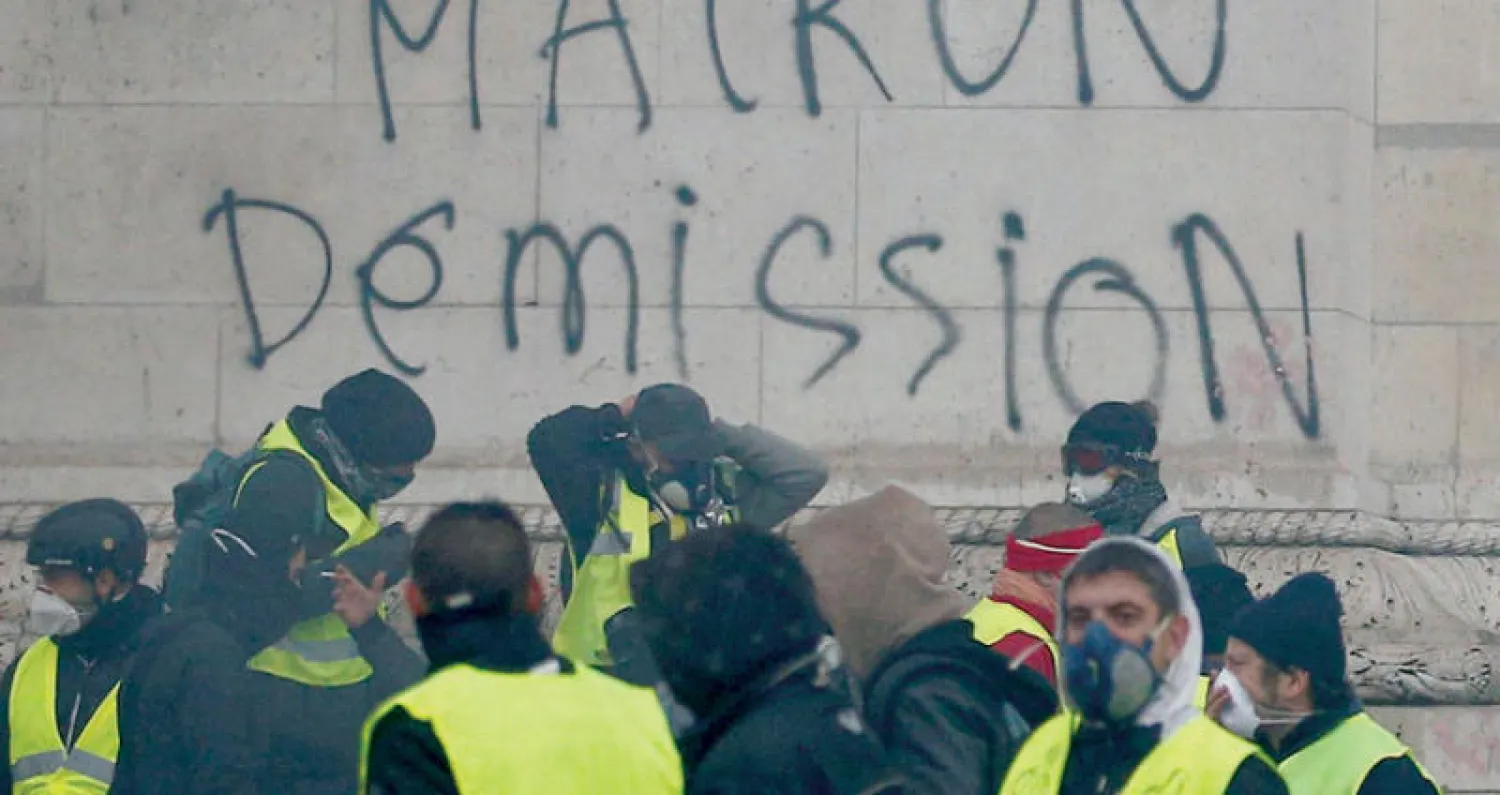 جانب من المحتجين بستراتهم الصفراء يقفون أمام حائط وسط شوارع باريس كتبت عليه عبارة «ماكرون استقِل» (رويترز)