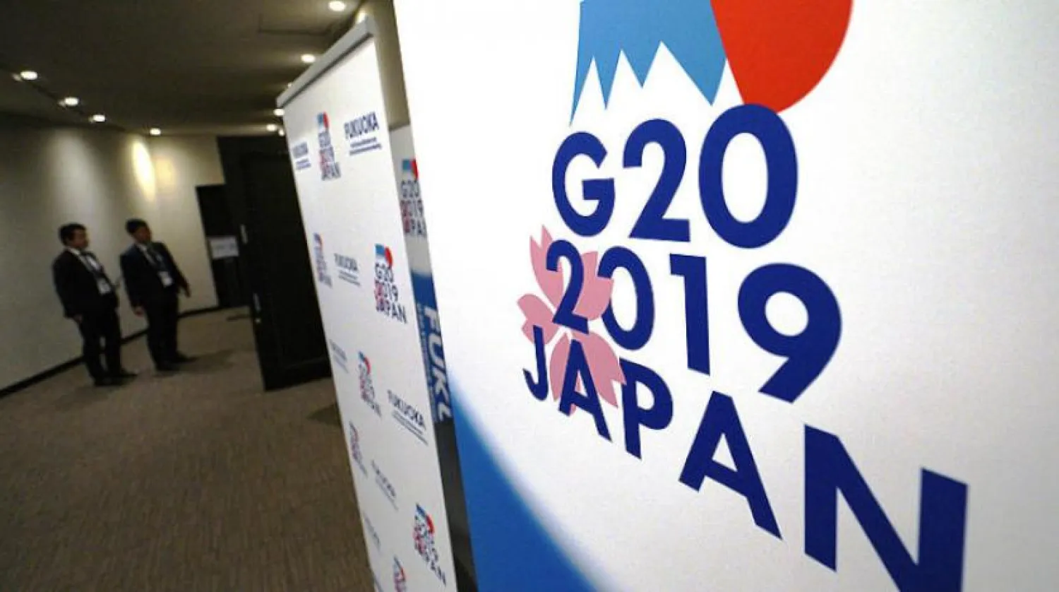 Japonya’nın batısındaki Fukuoka şehrinde bulunan basın merkezinin girişinde, G20 logosu önünde duran görevliler (AP)
