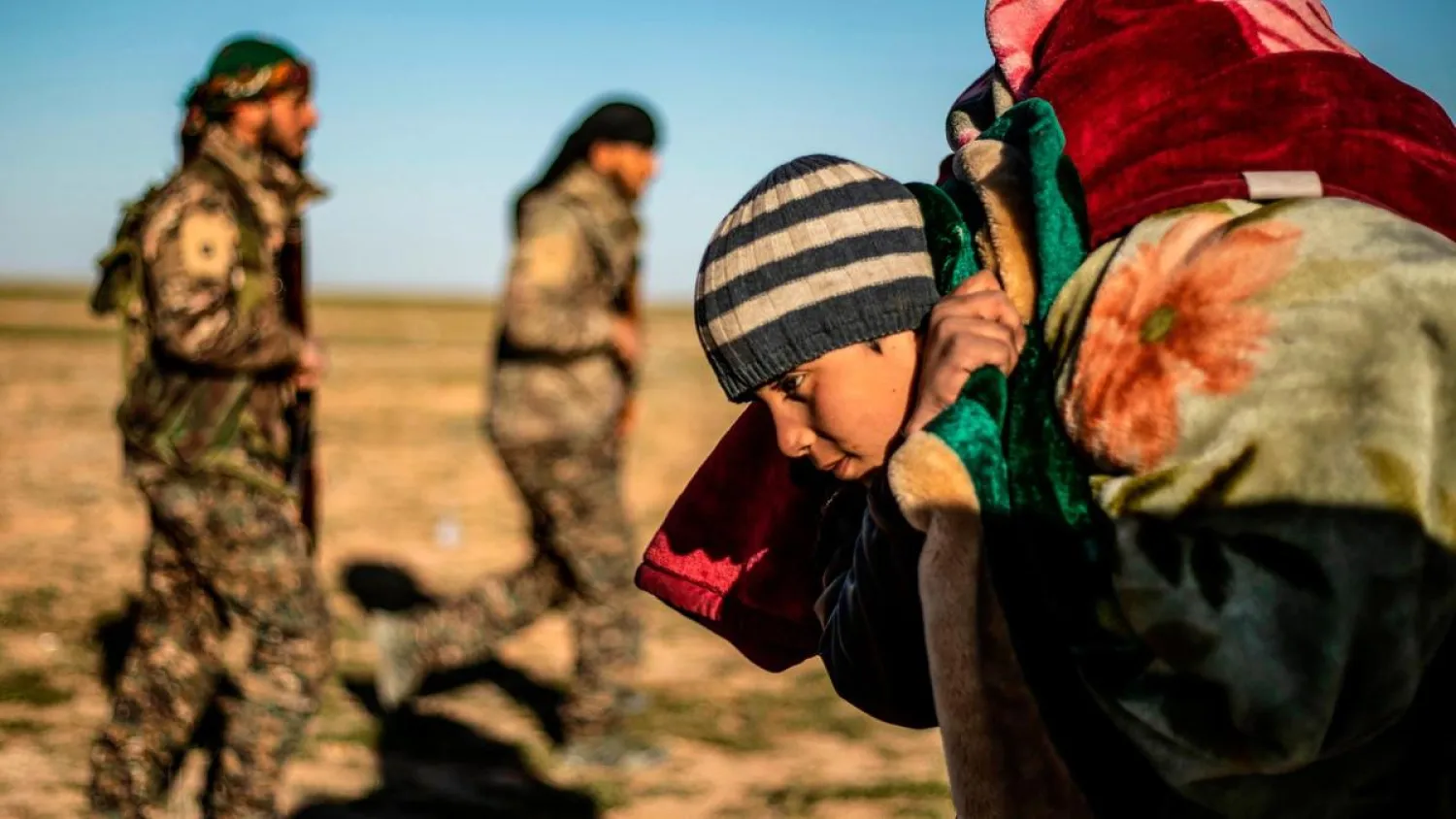 A child carrying covers on his back walks past members of the Kurdish-led Syrian Democratic Forces after leaving Baghouz. (AFP)