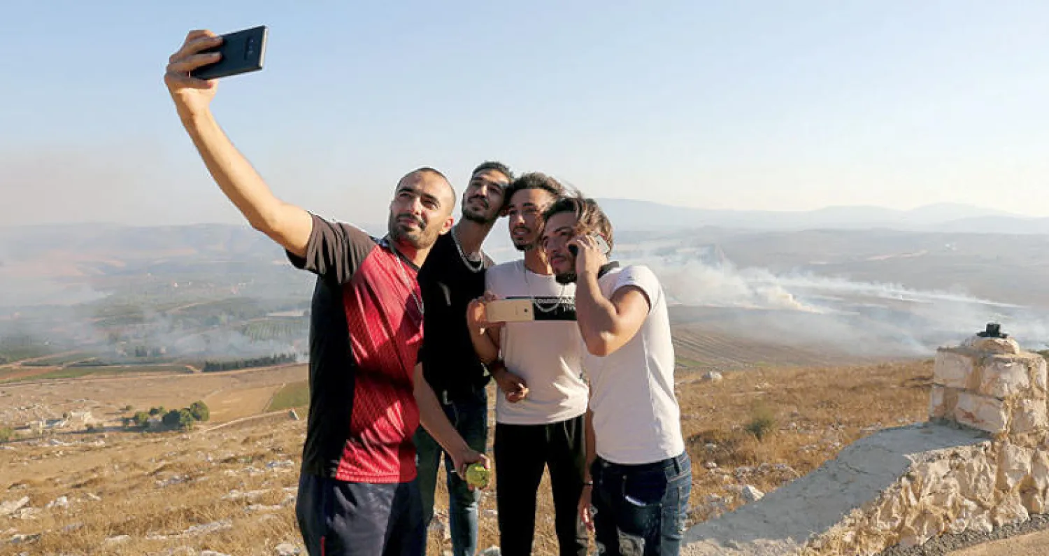 يلتقطون (سيلفي) وخلفهم دخان حرائق جراء قصف إسرائيلي على قرية مارون الراس (رويترز)