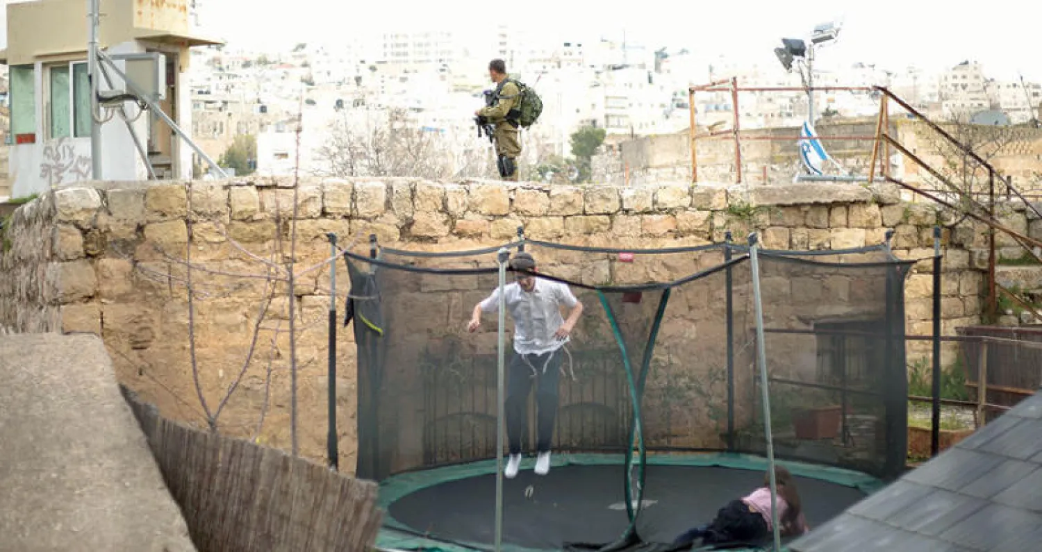 جندي اسرائيلي يحرس مستوطنين يلهون على الترمبولين في الخليل بالضفة الغربية (أ.ب)