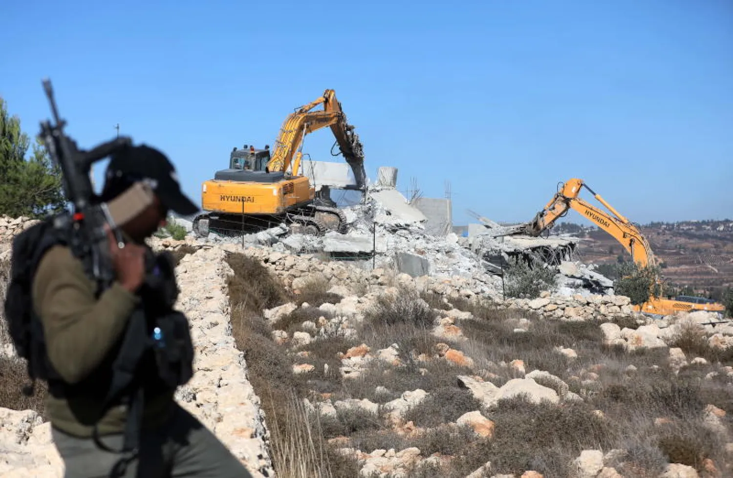 جندي إسرائيلي أمام جرافات تهدم منازل بالقرب من مخيم العروب للاجئين شمال مدينة الخليل بالضفة الغربية (إ.ب.أ)