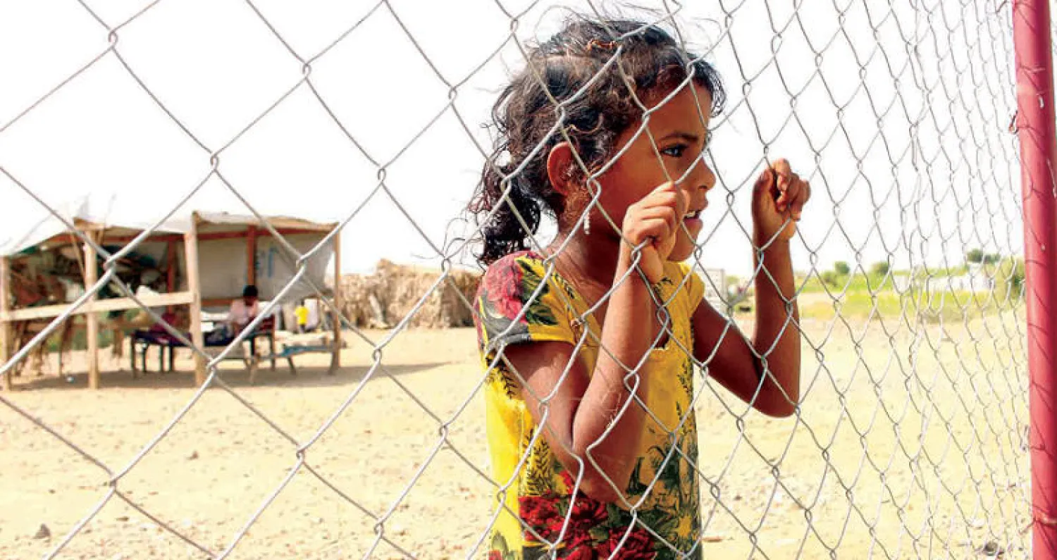 طفلة نازحة تنظر عبر سياج داخل مخيم في مديرية عبس التابعة لمحافظة حجة اليمنية (رويترز)