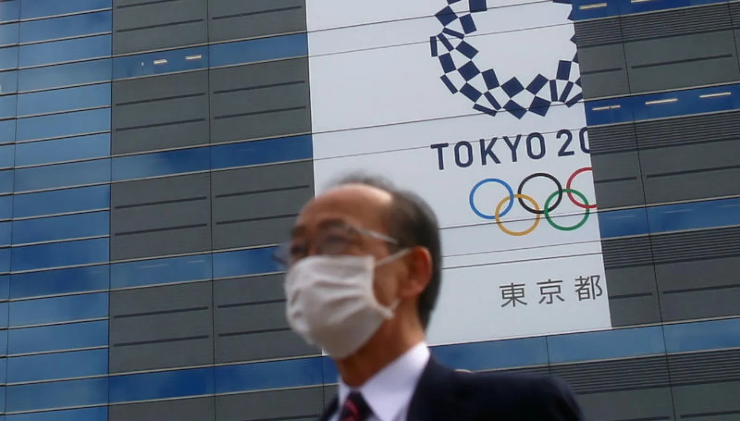 رجل يرتدي قناعاً واقياً أمام لوحة إعلانية لشعار أولمبياد طوكيو 2020 (رويترز)
