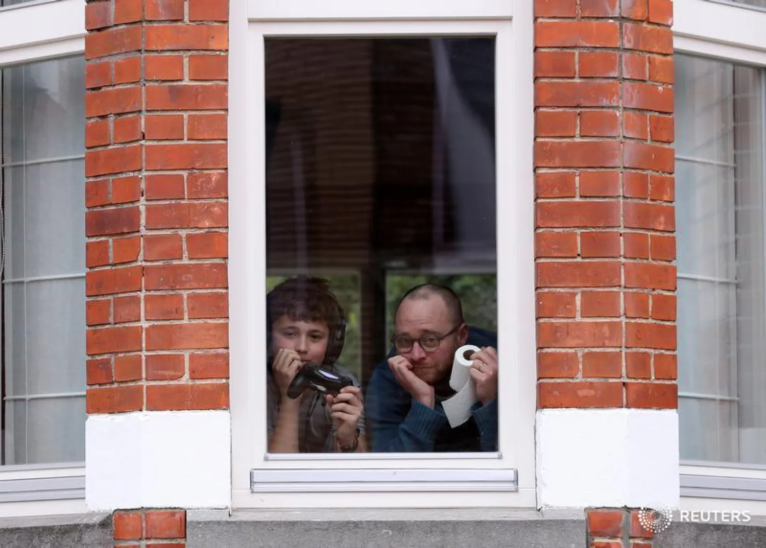 Resident Laurent Lanthier and his son Robin, 12 pose behind the window of their home with objects significant to them during a coronavirus lockdown in Brussels, Belgium. | Yves Herman/ REUTERS