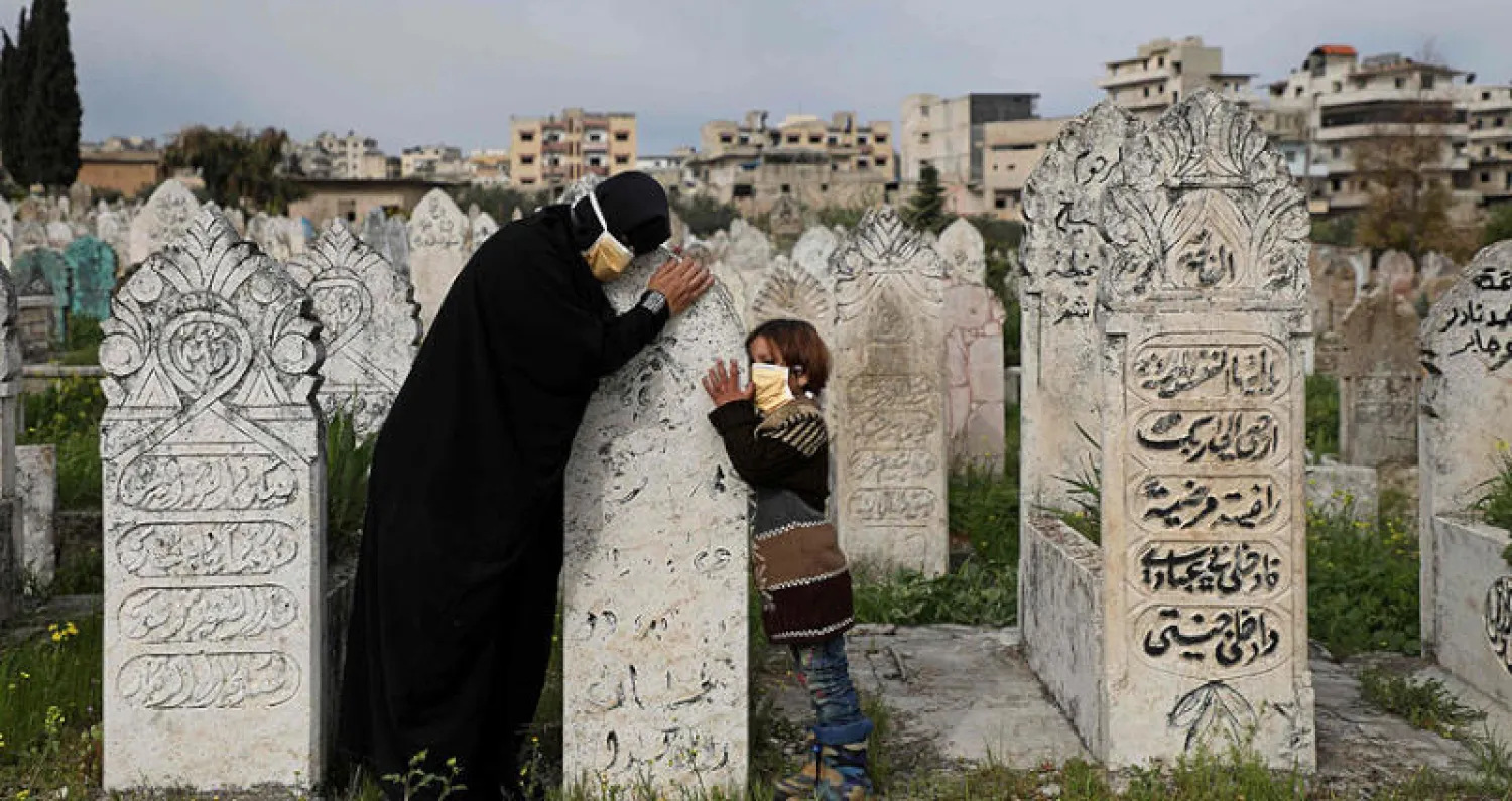 نازحة وابنتها ترتديان كمامتين واقيتين من {كورونا} تزوران قبر قريب لهما في أريحا بريف إدلب شمال غربي سوريا أمس (أ.ف.ب)