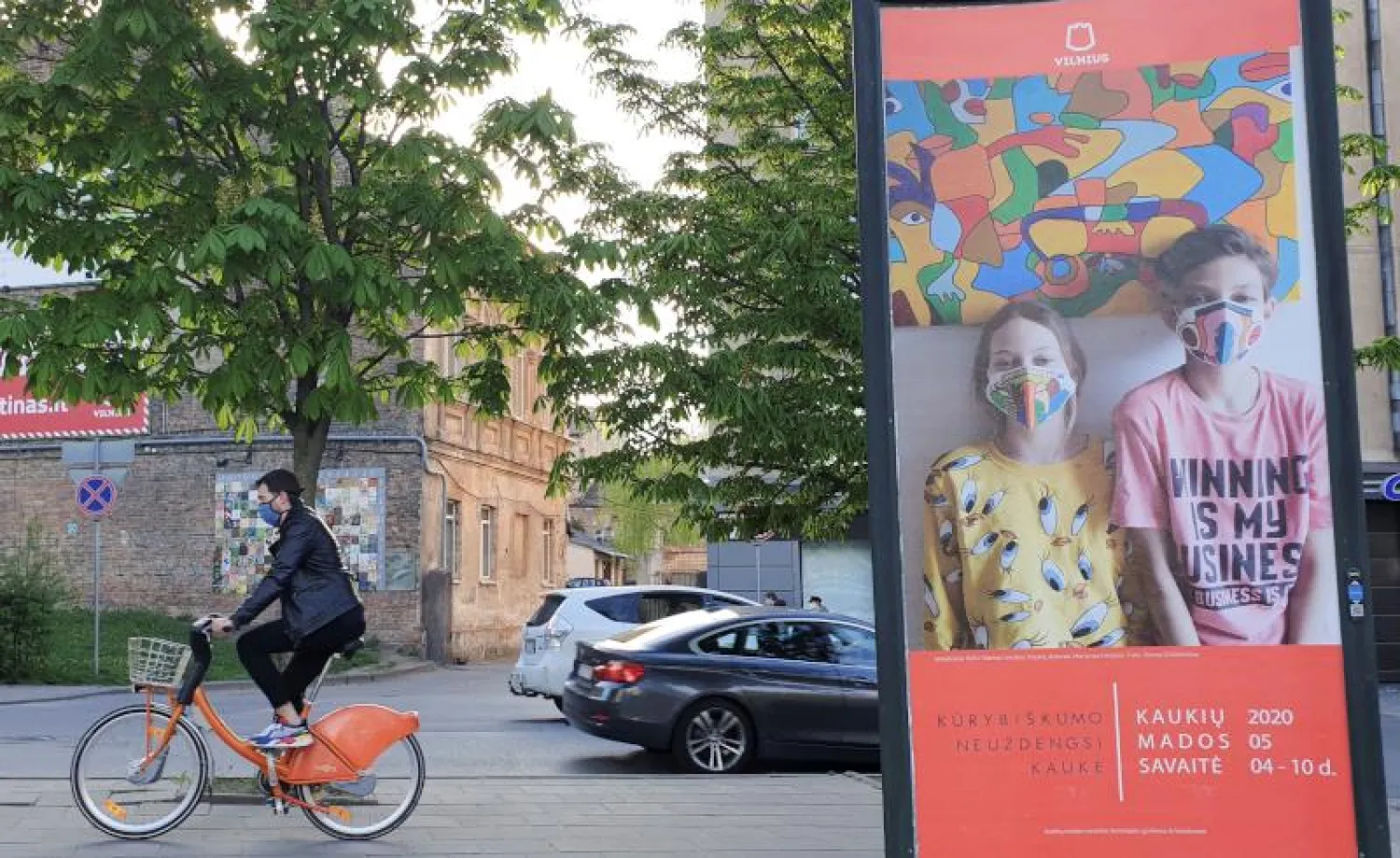 سائق دراجة هوائية يمر إلى جانب لوحة إعلانات تُظهر طفلين يرتديان قناعين واقيين ضمن أسبوع موضة الكمامات في ليتوانيا (رويترز)