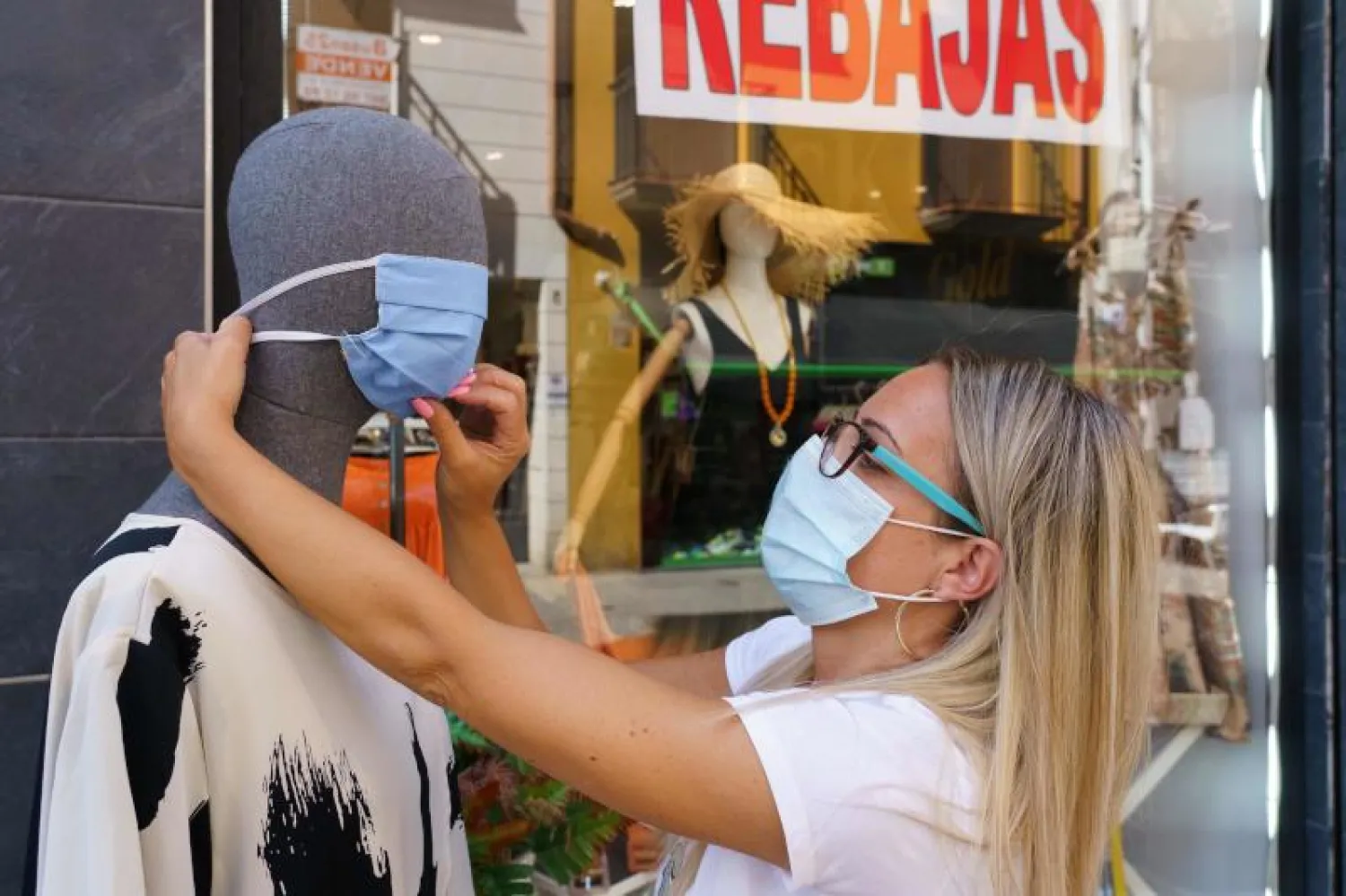 امرأة ترتدي كمامة وسط جائحة كورونا وتضع قناعاً على مجسم خارج متجر في إسبانيا (أ.ف.ب)