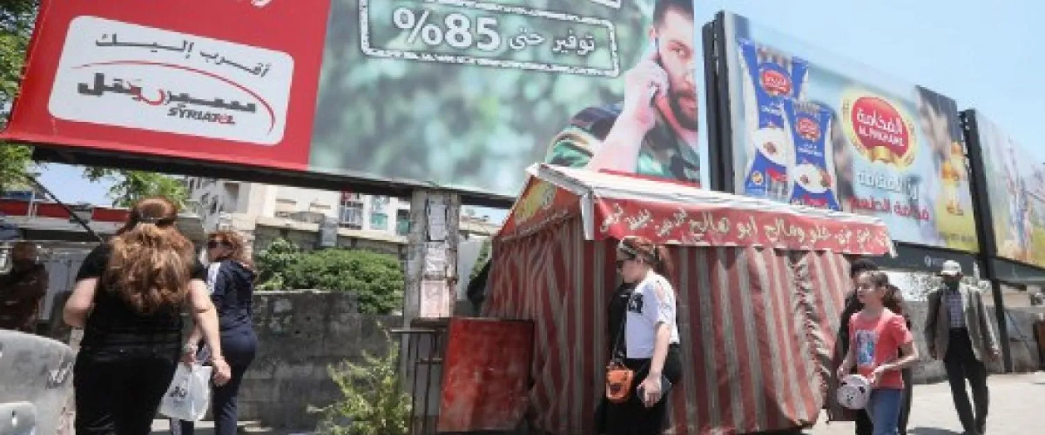 People walk underneath an advertising billboard of Syria's largest mobile operator Syriatel, owned by businessman Rami Makhlouf, in Damascus, 11/5/2020 (AFP)