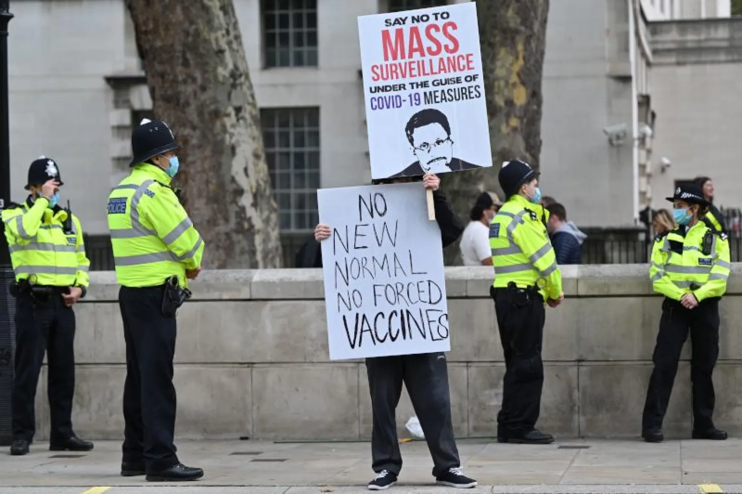 شرطيون يقفون في حراسة بينما يحمل متظاهر ملصقات في احتجاج ضد القيود المفروضة وسط لندن (أ.ف.ب)