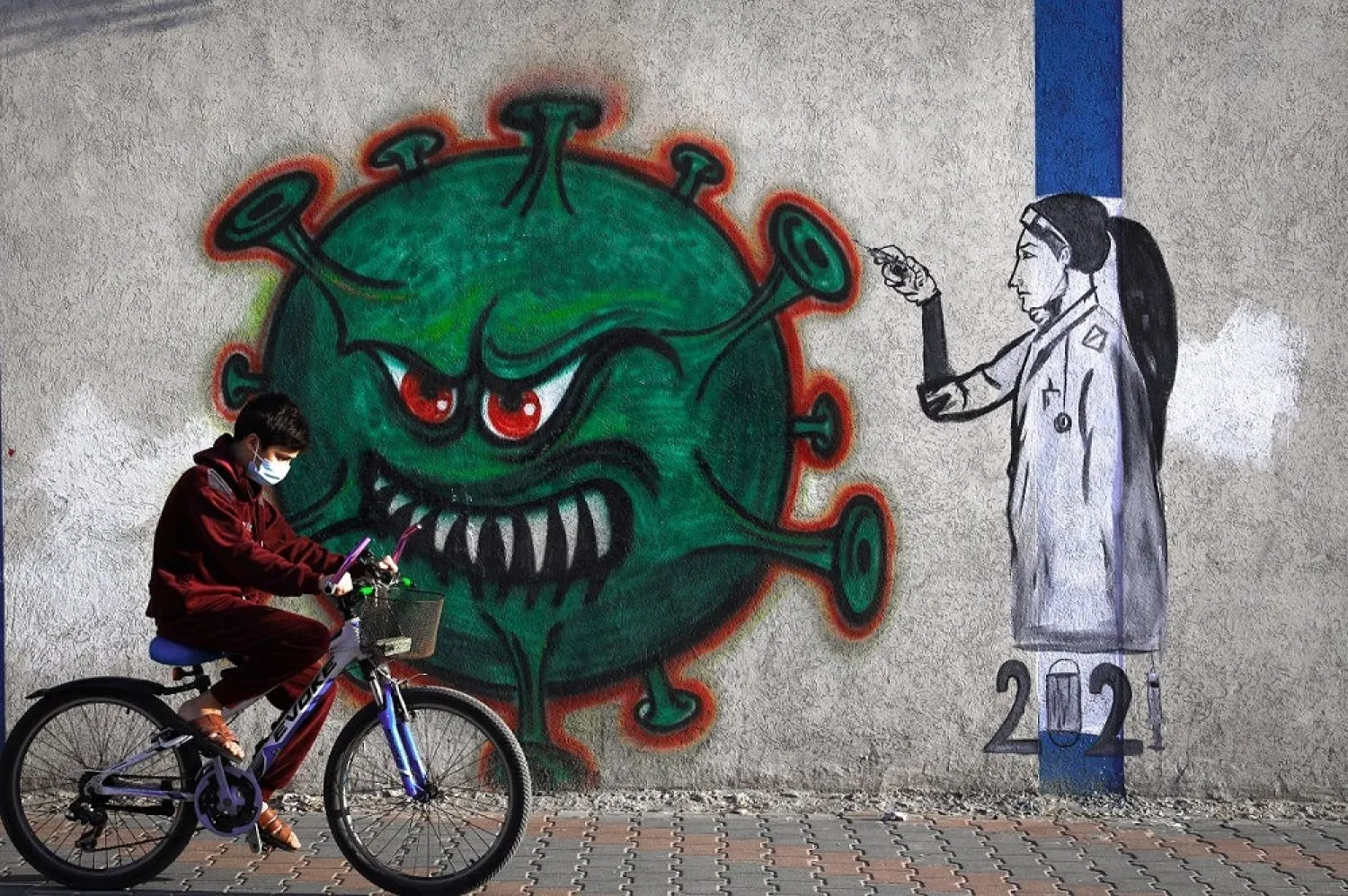 A Palestinian youth wearing a face mask rides his bicycle past a mural painting of a nurse injecting a vaccine to a COVID-19 virus in Gaza City, on December 31, 2020. (AFP)