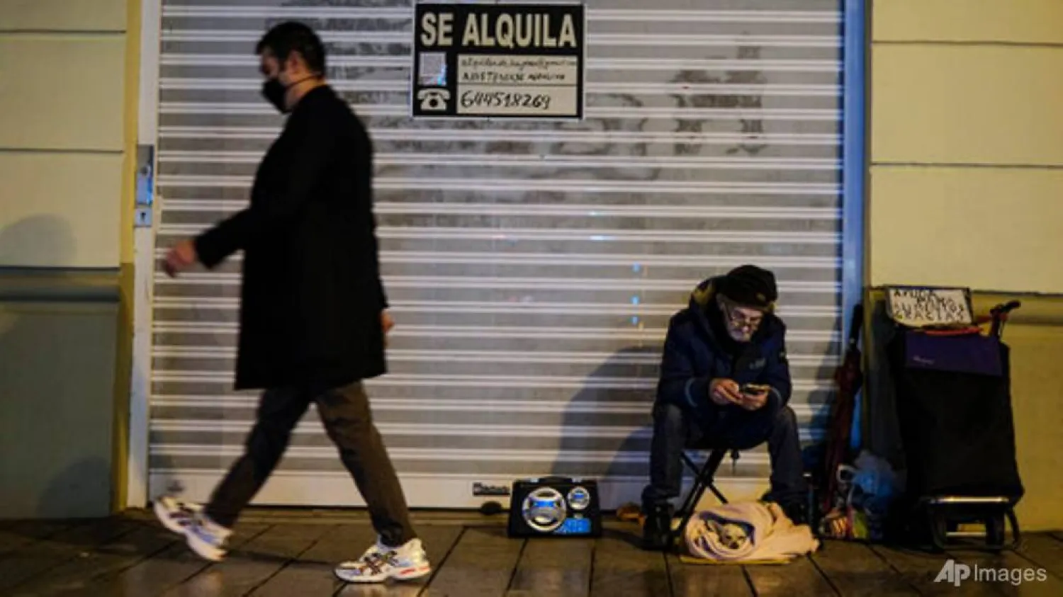 A homeless with his little pet, bottom right, in front a store to rent for food, begs for alms while pedestrian walking past wearing face mask protection against the coronavirus, in Pamplona, northern Spain, Thursday, Jan. 21, 2021. (AP Photo/Alvaro Barrientos)