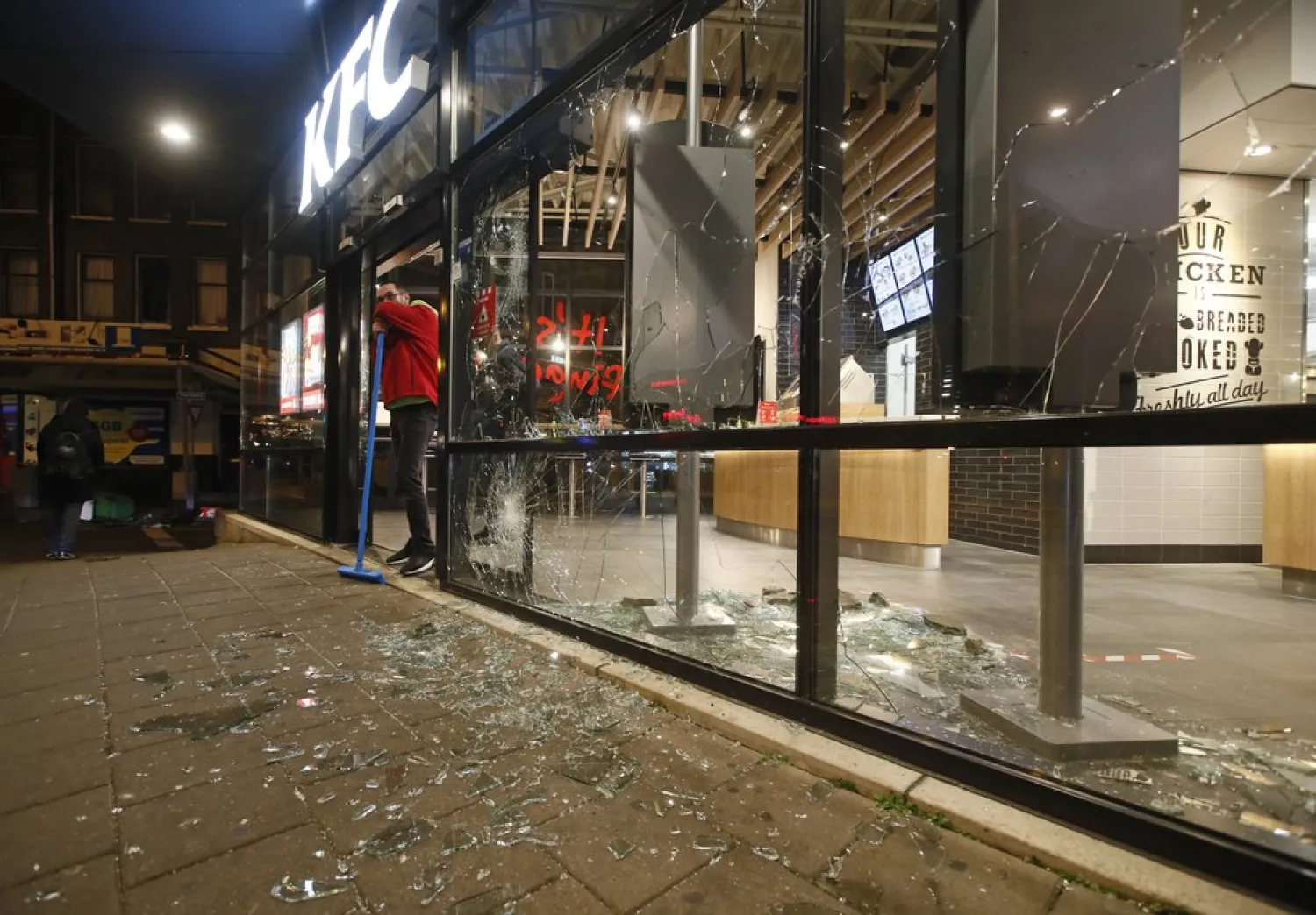 A man rests on his broom as he stands next to shards of glass and smashed windows of a fast-food restaurant that was damaged in protests against a nation-wide curfew in Rotterdam, Netherlands, Monday, Jan. 25, 2021. (AP)
