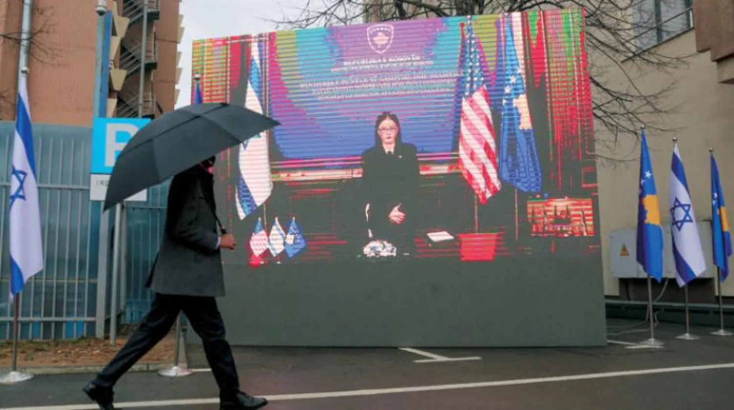 A government official holding an umbrella passes by a monitor displaying Kosovo's Foreign Minister Meliza Haradinaj-Stublla during a signing ceremony held virtually, in the capital Pristina, Monday, Feb. 1, 2021. (AP Photo/Visar Kryeziu)
