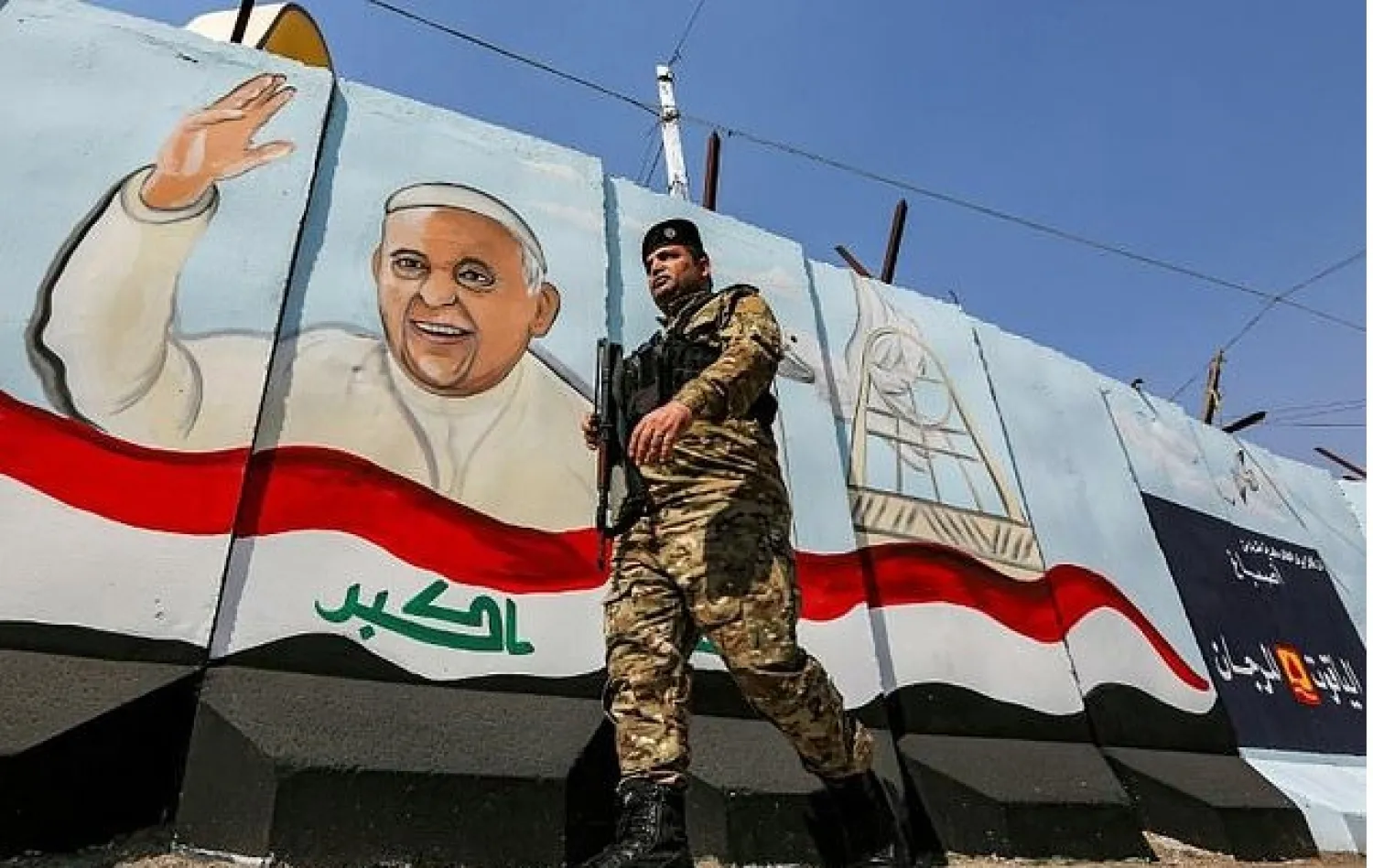 A member of the Iraqi security forces walks past a mural of Pope Francis with an Iraqi national flag, painted on a blast wall outside the Syriac Catholic Church of Our Lady of Deliverance in Baghdad - AFP

