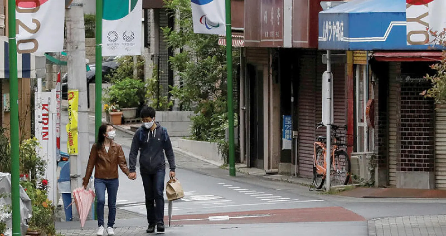يابانيان يسيران في أحد شوارع طوكيو بينما تبدو شعارات الأولمبياد المهدد بغياب الجماهير بسبب تفشي الوباء (الشرق الأوسط)