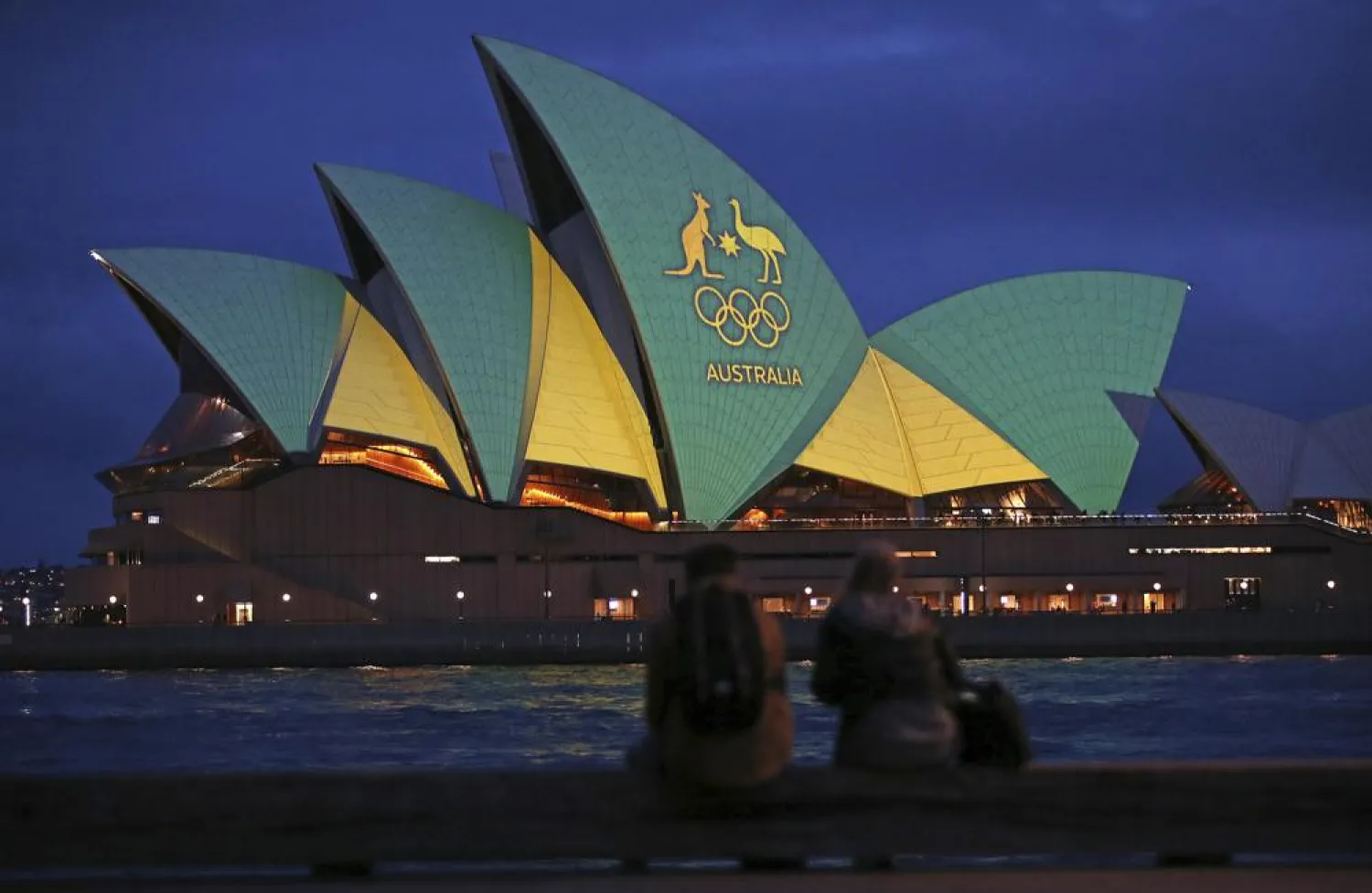 FILE - In this Friday, Aug. 5, 2016, file photo, a couple sit on a dock to look at the sails of the Sydney Opera House that are illuminated with the green and gold colors of the Australian Olympic team. (AP Photo/Rick Rycroft, File)
