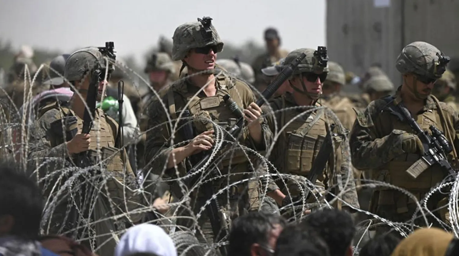 Afganlar Kabil Havaalanı’nın askeri bölümünün yakınında bir yolun kenarında otururken dikenli tellerin arkasından onlara bakan ABD askerleri (AFP)