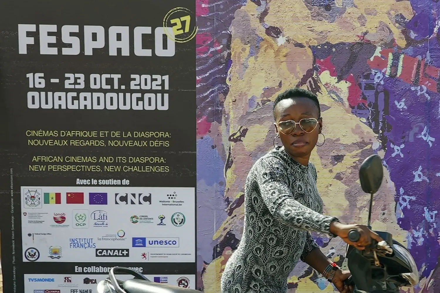 A woman pushes her scooter past the entrance of FESPACO (Pan-African Film & TV Festival of Ouagadougou) in Ouagadougou, Burkina Faso, Friday Oct. 15, 2021. (AP)