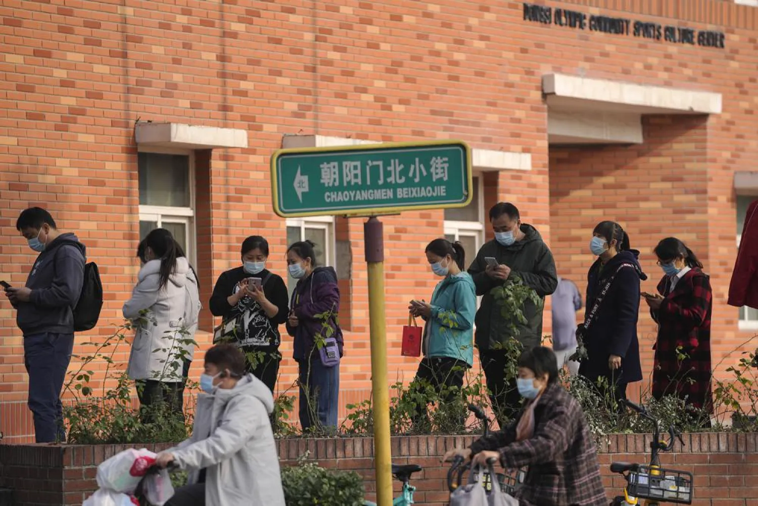 Women wearing face masks to help curb the spread of the coronavirus ride scooters passing by masked residents line up to receive booster shots against COVID-19 at a vaccination site in Beijing, Monday, Oct. 25, 2021.(AP Photo/Andy Wong)
