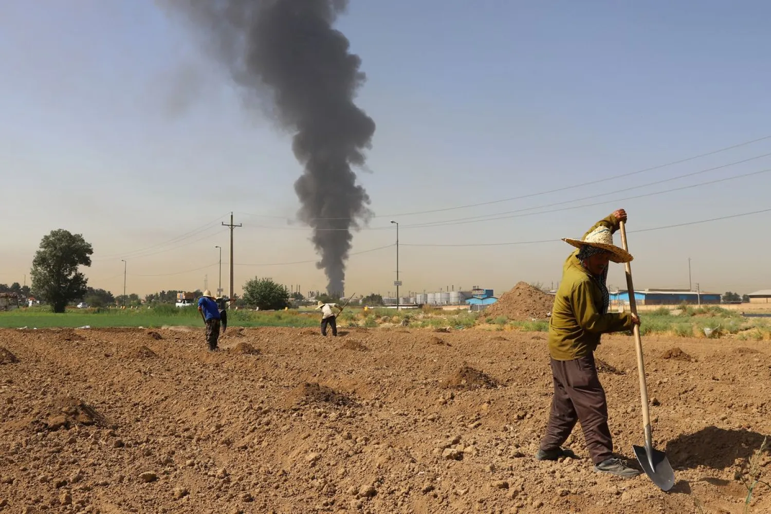 Farmers work in a field as smoke from an oil refinery rises in the background, in Tehran, Iran June 3, 2021. Majid Asgaripour/WANA (West Asia News Agency) via REUTERS/File Photo