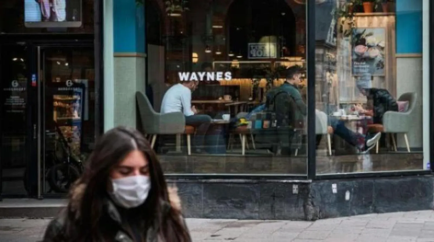 امرأة تسير وهي تضع كمامة وخلفها مقهى مزدحم بالزبائن في العاصمة السويدية استوكهولم (أ.ف.ب)