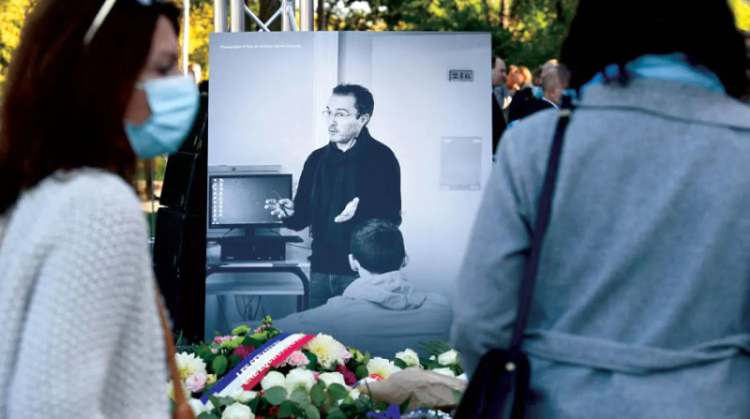 أفراد من الجمهور وأكاليل من الورود أمام صورة تصور مدرس التاريخ الفرنسي صمويل باتي أثناء حضورهم حفل تأبين لتكريمه شمال غرب باريس (أ.ف.ب)