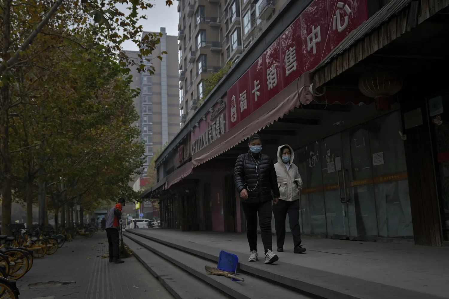Women wearing face masks walk by a worker sweeping along the shuttered shops as part of COVID-19 controls in Beijing, Thursday, Nov. 24, 2022. (AP)