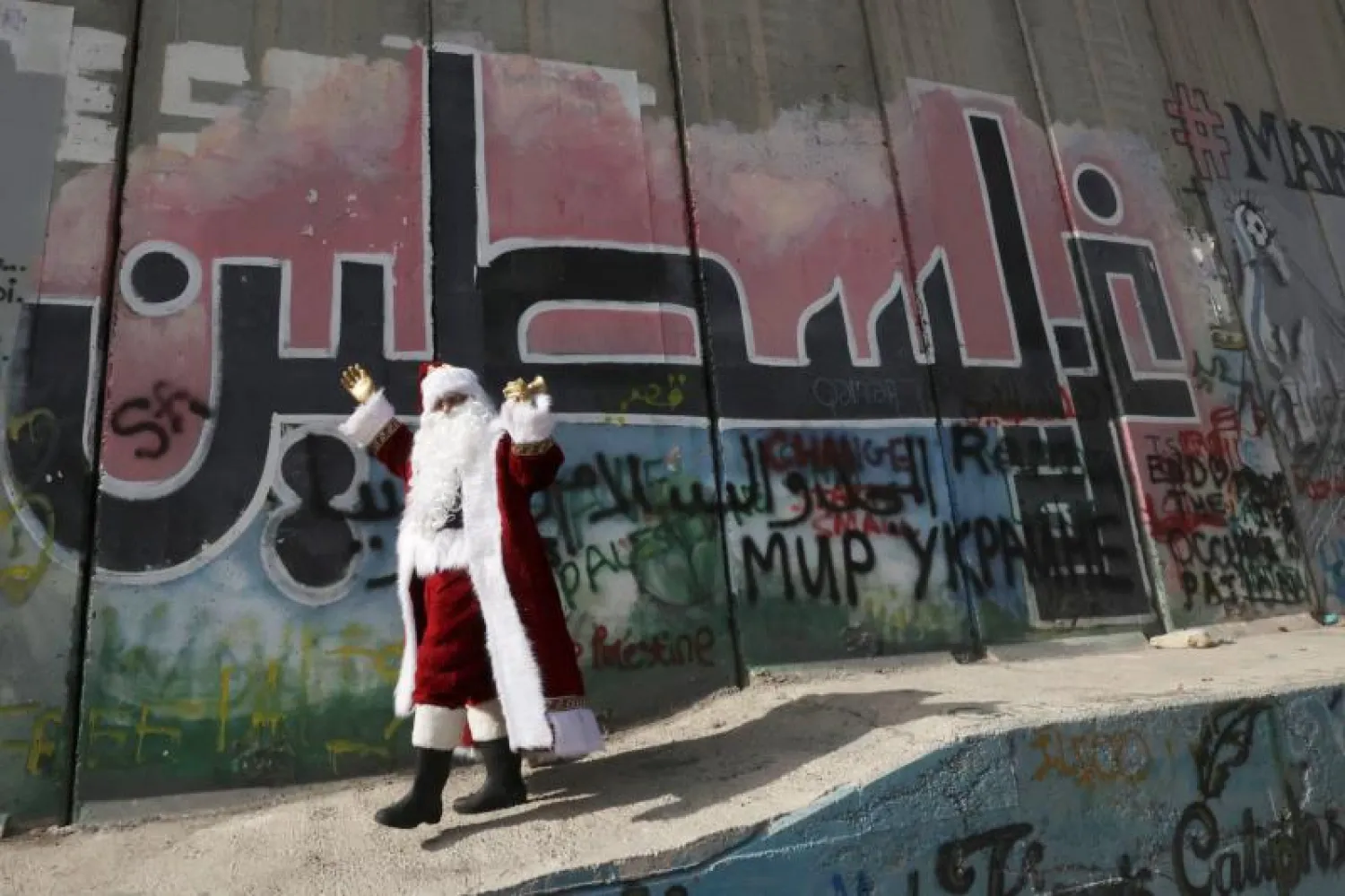 رجل يرتدي زي بابا نويل بجانب جدارية كتب عليها «فلسطين» على الجدار الفاصل في الضفة الغريبة (أ.ف.ب)