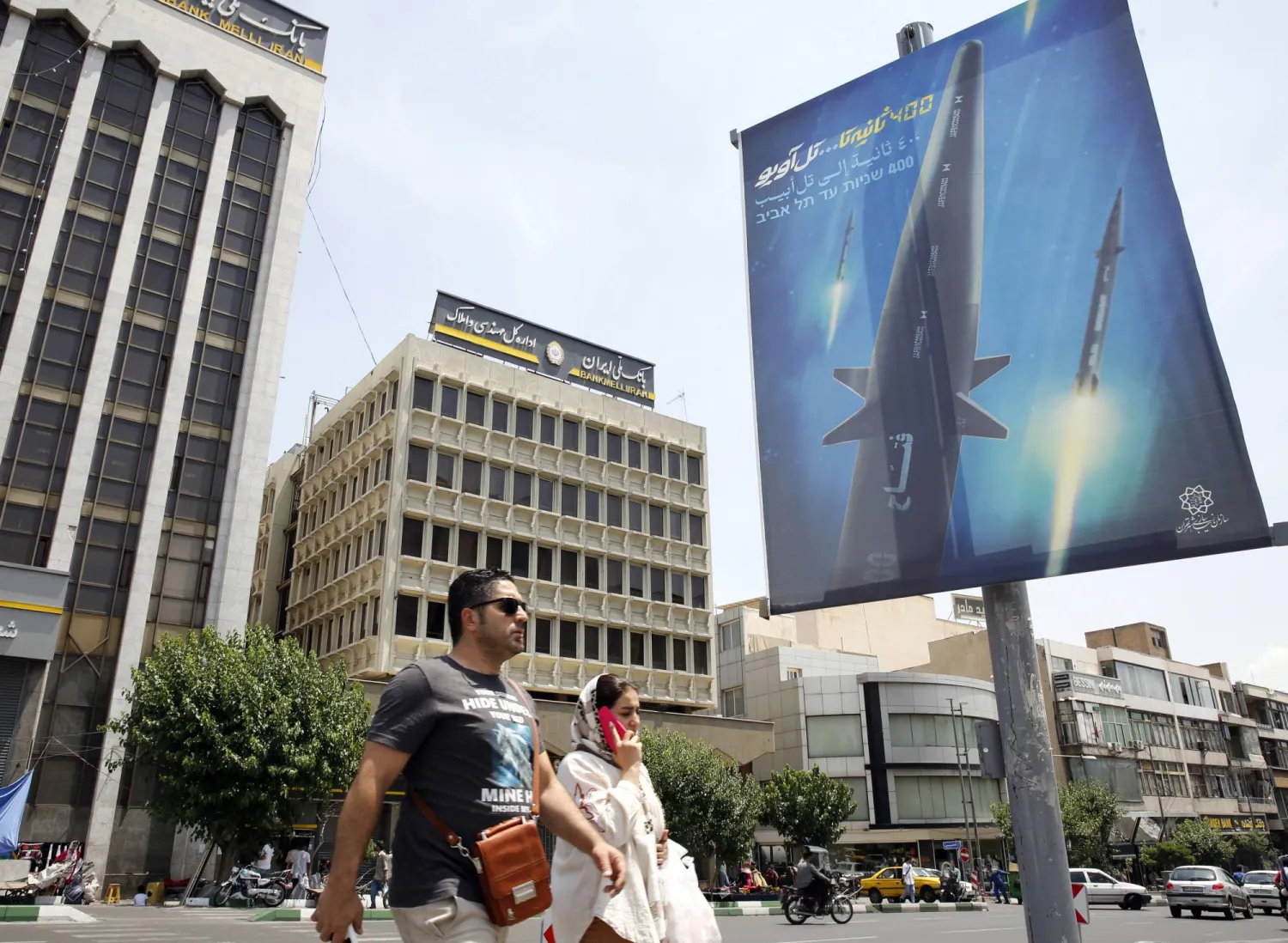 رجل وامرأة يمشيان بجوار لوحة دعائية لصاروخ «فرط صوتي» ويبدو شعار «400 ثانية إلى تل أبيب» في شارع وسط طهران (إ.ب.أ)