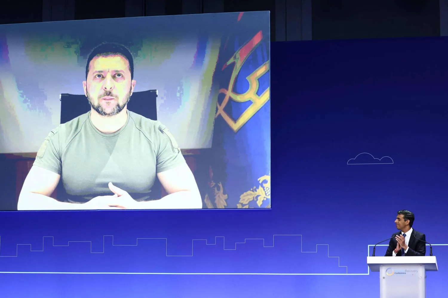 رئيس الوزراء البريطاني ريشي سوناك يصفق لرئيس أوكرانيا فولوديمير زيلينسكي على الشاشة في الجلسة الافتتاحية خلال اليوم الأول من مؤتمر «تعافي أوكرانيا» في لندن (أ.ف.ب)