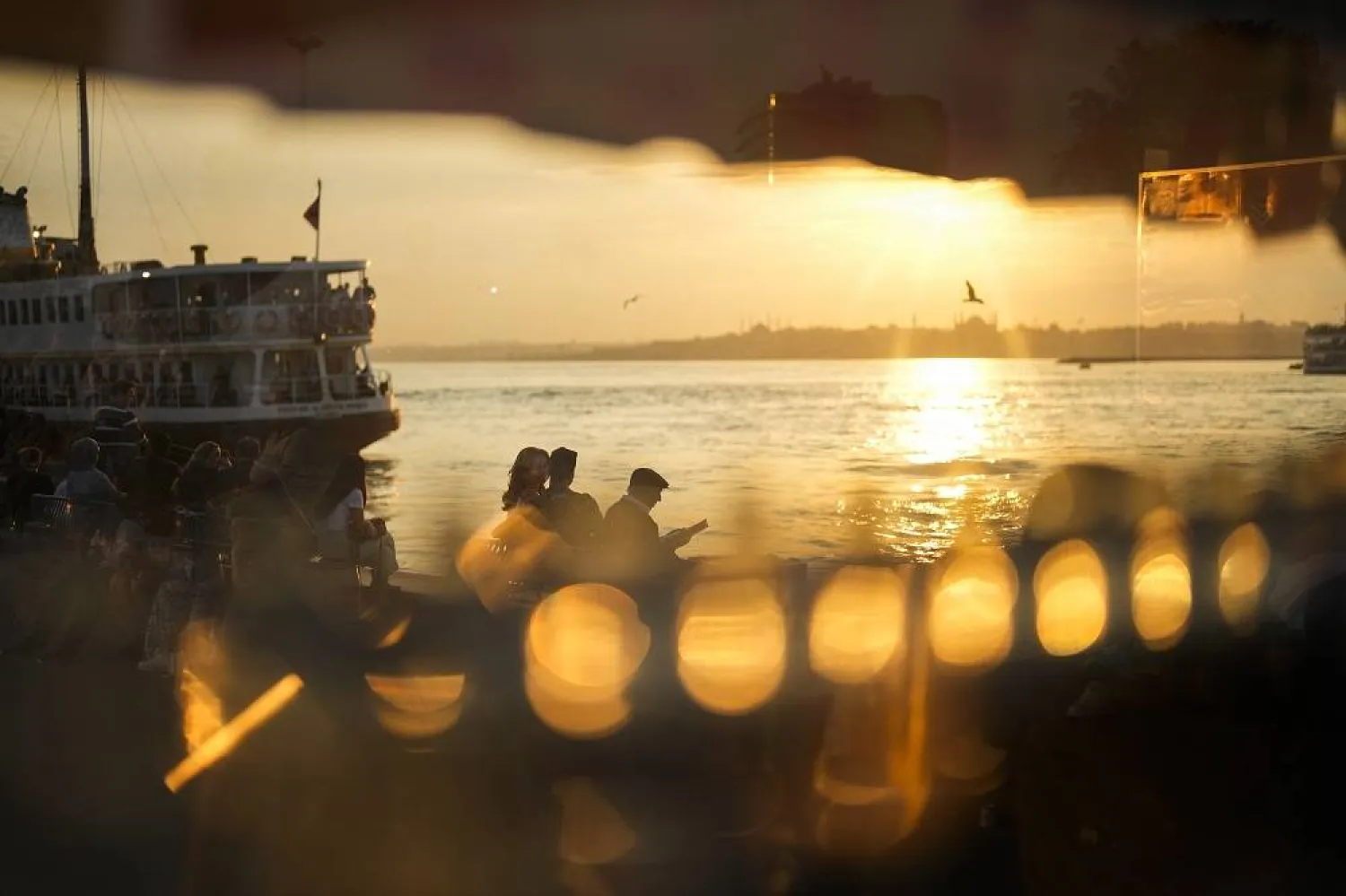People, seen through a glass of a street food stand, sit at Kadikoy sea promenade at the Bosphorus in Istanbul, Türkiye, Monday, June 19, 2023.  (AP)