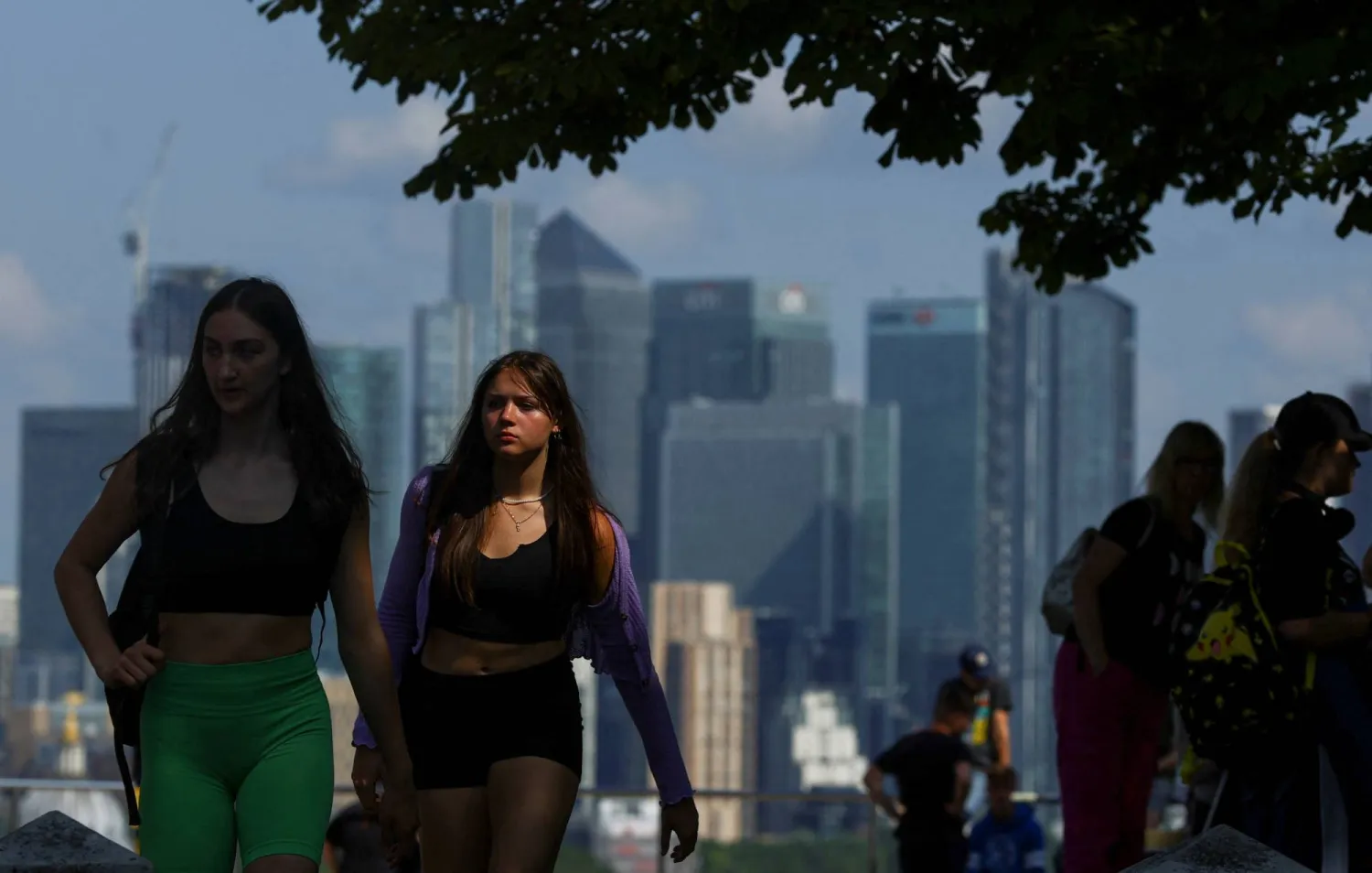 أشخاص يسيرون في غرينتش بارك وفي الخلفية الحي المالي لمدينة لندن ببريطانيا (رويترز)